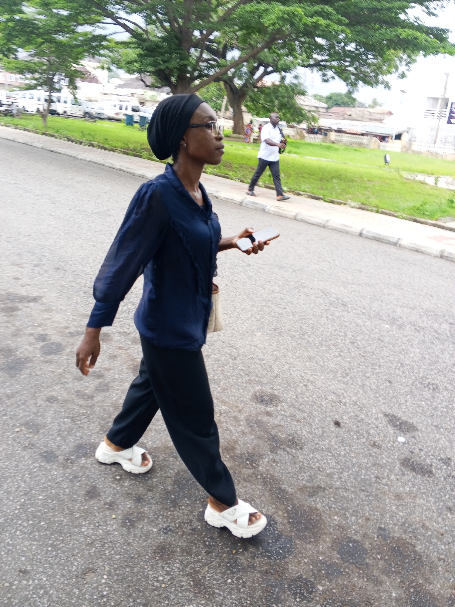 A full-body, outdoor, eye-level, medium shot captured on an overcast day shows a young Black woman walking on a paved street in Lokoja, Nigeria. She is wearing a dark blue long-sleeved shirt, dark pants, and white sandals. She has a black headscarf and glasses on, and she is holding a smartphone in her right hand. She is walking to the right of the frame. In the background, a man in a white shirt and dark pants walks to the left. Further in the distance, white vans are parked in front of buildings. Trees with green foliage line the street. The ground is paved with asphalt, and there is a sidewalk to the right.