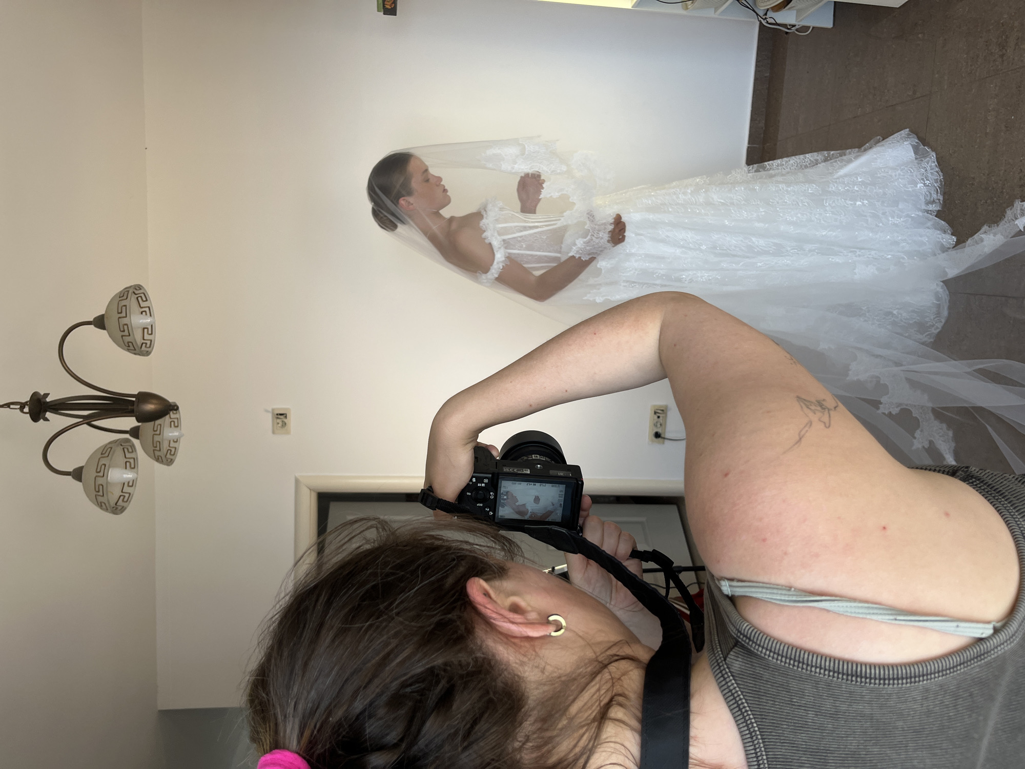A woman in a grey tank top photographs a bride in a white wedding dress with a veil. The photographer is in the foreground on the left, holding a camera to her eye. The bride is in the middle ground on the right, facing left and looking down. She has her hair up in a bun. The wedding dress is off-the-shoulder with lace detailing and a long train. The veil is also long and has lace trim. The background is a plain white wall with a chandelier on the ceiling. There is a doorway on the left and electrical outlets on the right. The floor is tiled. The scene is indoors.