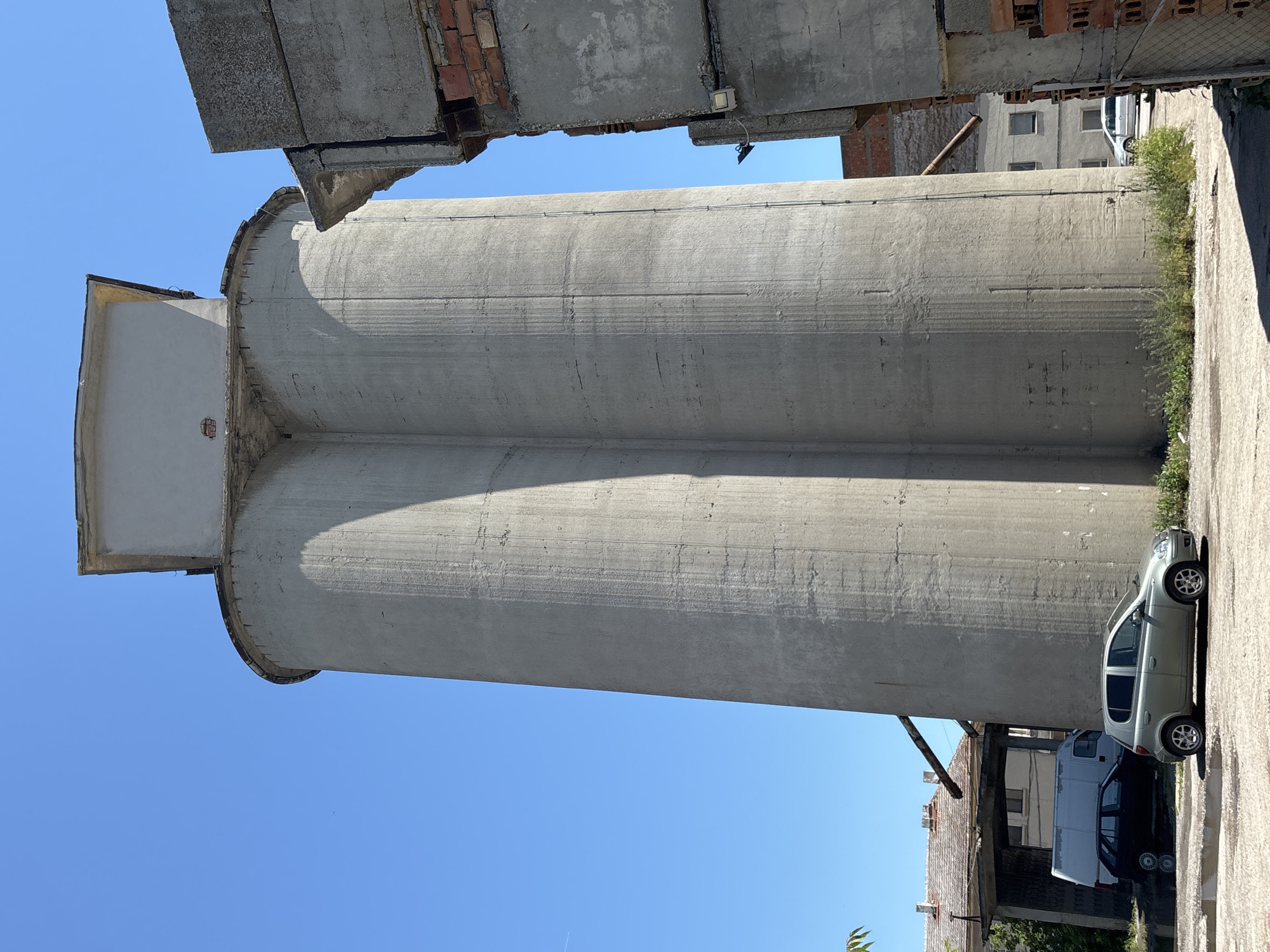 In Kavarna, Bulgaria, a towering concrete structure, likely a silo, dominates the frame under a clear blue sky. The silo appears to be comprised of two connected cylindrical sections, with a rectangular cap at its apex. Its weathered concrete surface bears vertical lines and subtle cracks. To the right, a partially constructed or dilapidated concrete and brick building stands, its exposed rebar and brickwork suggesting ongoing work or decay. In the foreground, a silver hatchback car is parked on a paved surface, with a black hatchback and a white van visible behind it. Vegetation sprouts around the base of the silo and the adjacent buildings, indicating an outdoor, potentially industrial or semi-abandoned, setting. The scene is illuminated by bright sunlight, casting distinct shadows.