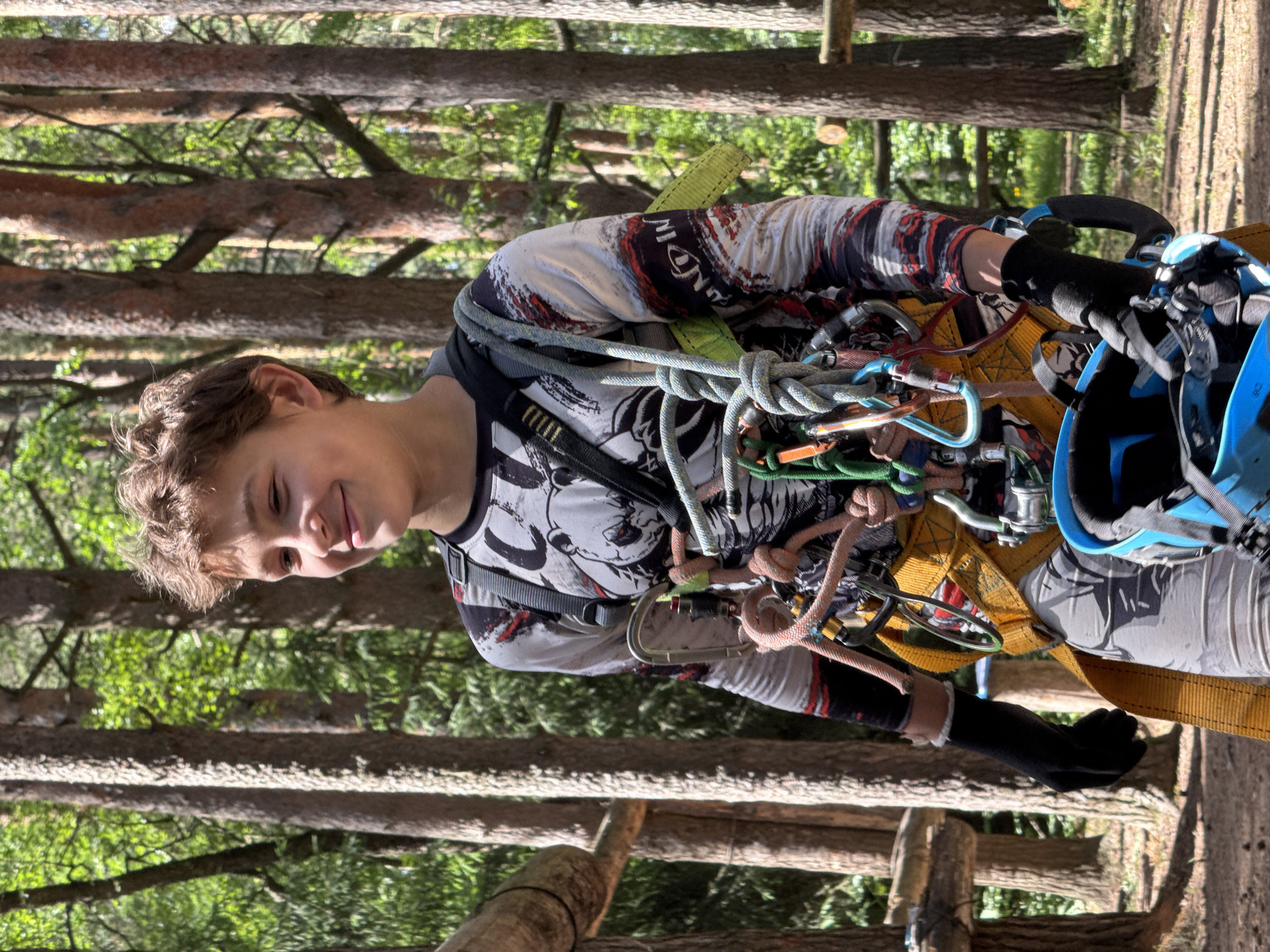 This media file shows a young boy in an adventure park setting, likely participating in a ropes course or zip-lining activity. He is wearing a grey and white long-sleeved shirt with a graphic print, a climbing harness, and a blue helmet. The harness is equipped with multiple ropes and carabiners, indicating preparation for an activity that requires safety equipment.
The boy is smiling and looking towards the camera, suggesting he is enjoying himself and feeling confident. His expression conveys a sense of excitement and fun. The background is a forest with tall trees and dappled sunlight, implying it is daytime and the weather is pleasant. The presence of ropes and a harness, along with the natural wooded environment, strongly suggests the location is an outdoor adventure park or a similar recreational facility in Chaikovsky, Russia.
Notable details include the intricate arrangement of the climbing gear and the vibrant green foliage of the trees. The sunlight filtering through the leaves creates a bright and airy atmosphere. The print on the boy's shirt includes some visible text, but it is difficult to read clearly due to the resolution and angle. However, there appears to be a stylized logo or design with letters, possibly "MONDAY" or similar, rendered in a black and red color scheme. The overall scene depicts a child actively engaged in an outdoor adventure, experiencing the thrill of an activity amidst a natural setting.