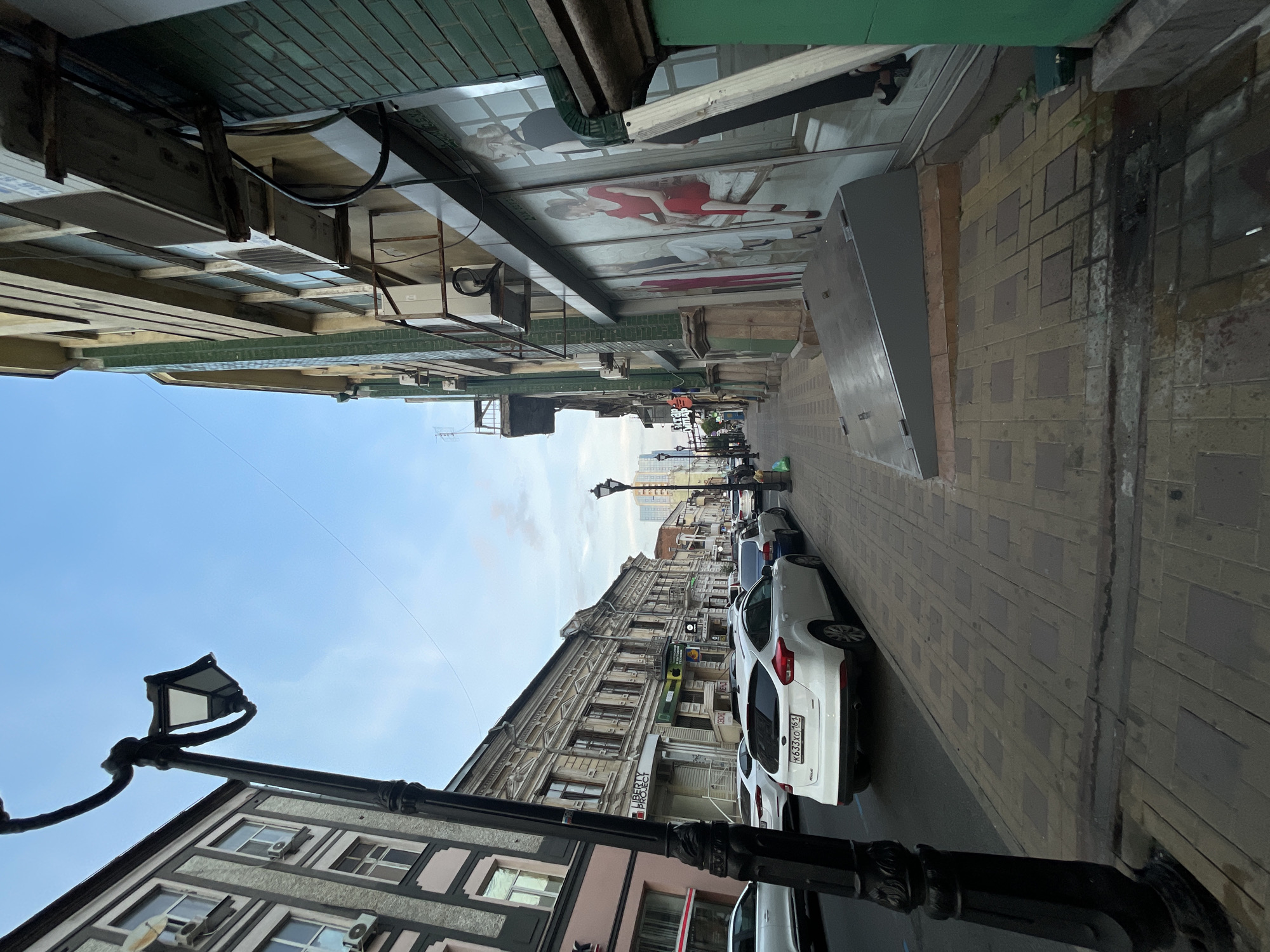 The image depicts a street scene in Rostov-on-Don, Russia. A sidewalk paved with interlocking bricks runs along the right side of the street, bordered by buildings. On the left side of the street, there are parked cars, including a white Ford Focus with a license plate K633XO 161. The buildings lining the street have architectural details characteristic of older European city centers. One building on the right features large glass windows with images of women displayed. A black ornate lamppost stands prominently in the foreground on the left. The sky is visible in the background, appearing partly cloudy and blue. The scene appears to be during daylight hours, likely late afternoon or early evening, given the lighting.