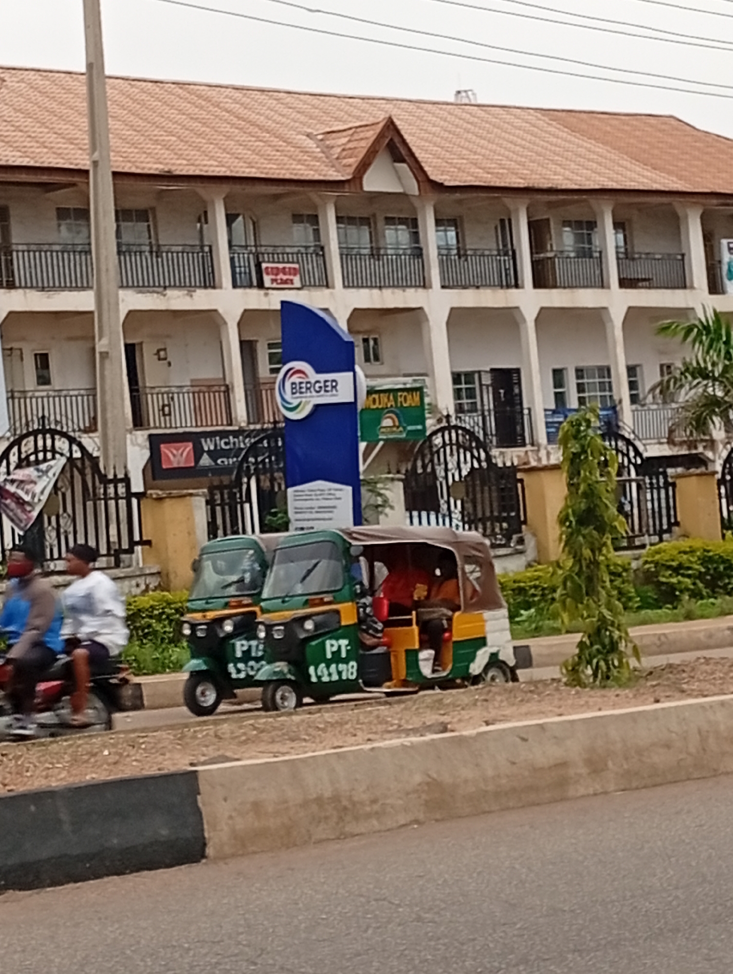 The image depicts a street scene in Gura Top, Nigeria, characterized by a two-story building with multiple windows and balconies, topped with a tiled roof. In front of the building, there are several vehicles and structures. Two auto-rickshaws, also known as Keke NAPEPs, are parked on the side of the road. The auto-rickshaws are green and yellow, with one displaying the license plate "PT 1302" and the other "PT 14178". A motorcycle is also visible, carrying two individuals, one wearing a blue shirt and the other a white shirt. A large blue sign with the "BERGER" logo and some text in a smaller font is positioned near the road. Adjacent to the Berger sign, another sign reads "MOUKA FOAM". A dark metal gate with ornate designs is situated in front of the building. The environment appears to be a commercial or residential area, with a concrete road and sidewalk in the foreground. Lush green bushes and a small tree are present along the side of the road. The sky is overcast.