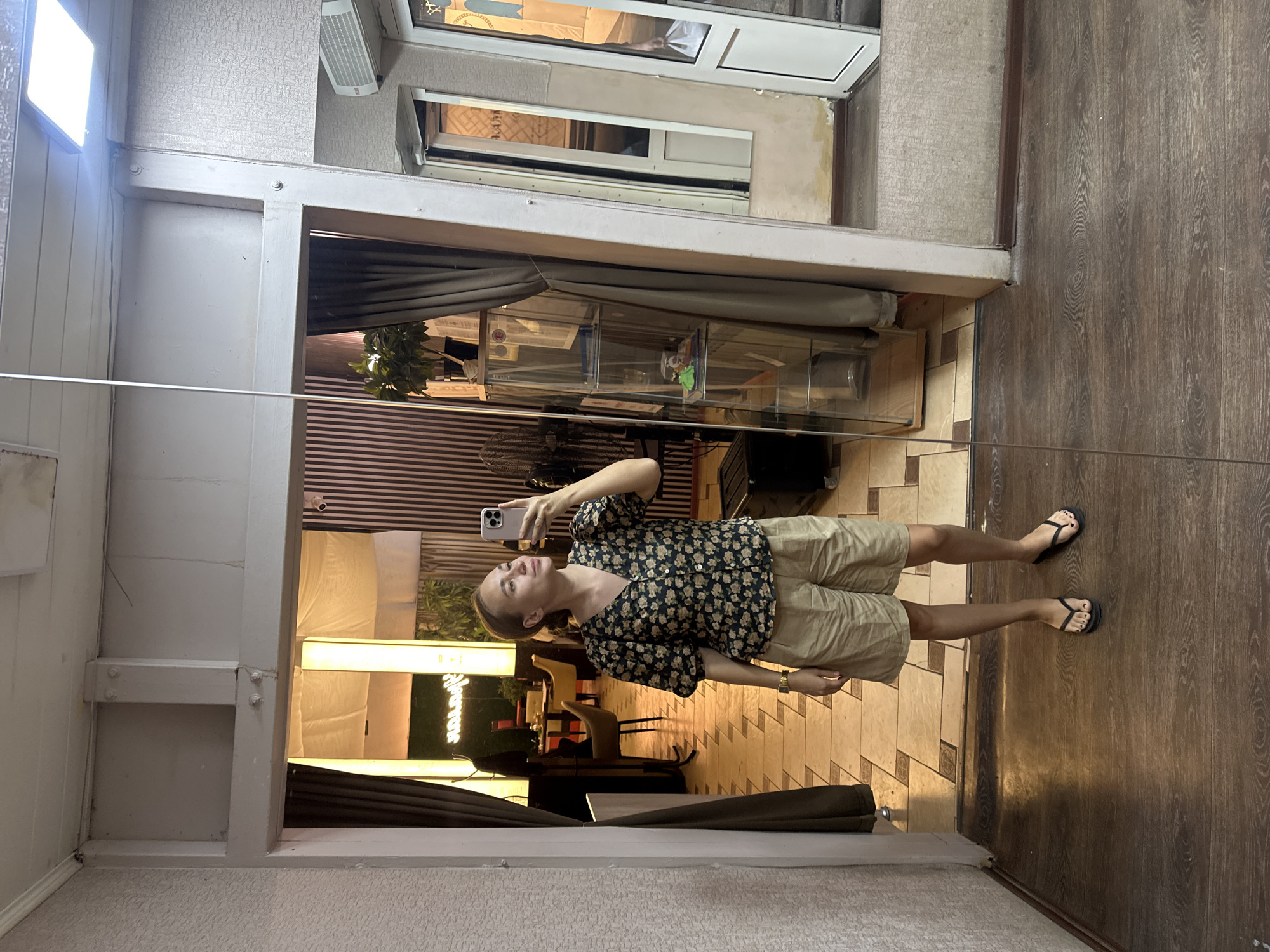 A woman is taking a mirror selfie in what appears to be a commercial establishment, possibly a salon or shop, in Elista, Russia. She is wearing a dark, floral-patterned short-sleeved shirt with puffed sleeves and beige, knee-length shorts. She has on black flip-flops and a gold watch on her left wrist. She is holding a smartphone in her right hand, taking the picture.
The background shows a hallway or interior space with reflective surfaces. To her left, through an open doorway, is a dining area with tables and chairs, illuminated by bright, possibly neon, signage. To her right, a large mirror reflects another part of the interior, including a glass display cabinet with various items and a doorway leading to an outdoor area. The flooring is a mix of wood-effect planks and patterned tiles. The walls are textured, and there are curtains and overhead lighting fixtures visible. The overall lighting suggests an indoor evening setting.