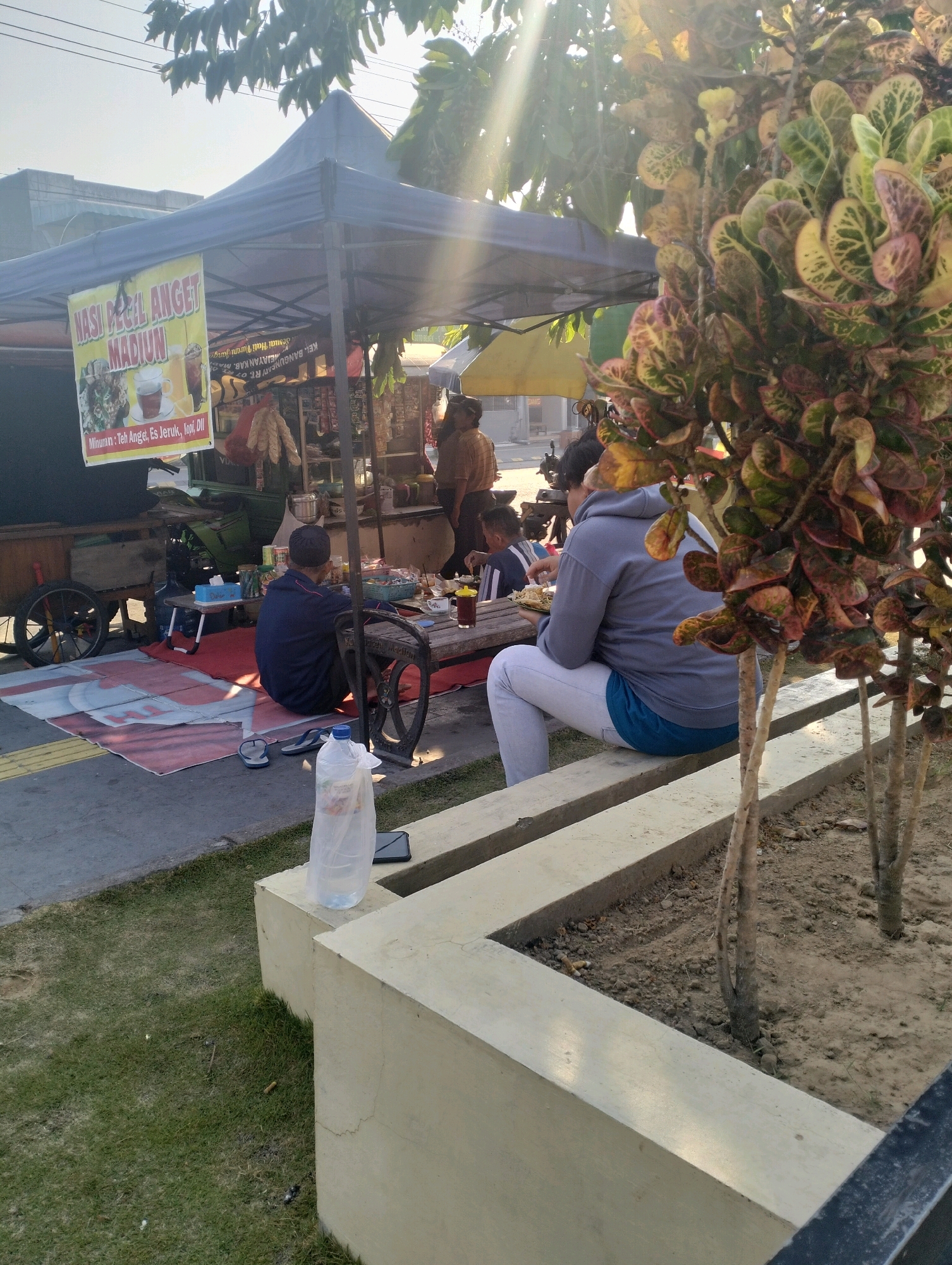 The image depicts an outdoor food stall in Bangunsari, Indonesia, bathed in bright sunlight filtering through trees. A blue canopy covers the stall, displaying a banner that reads "JESI PECEL ANGET MADIUN," advertising a local dish and drinks like "Teh Anget, Es Jeruk, Kopi, dll." A wooden cart with large wheels is parked to the left, and a green tarp is visible behind it.
In the foreground, two individuals are seated on a concrete bench. One, wearing a grey hoodie and white pants with a blue undershirt, is facing away from the camera, holding a plate of food. The other person, in a dark blue jacket and a dark head covering, is also seated, facing the stall. A red mat lies on the ground in front of the stall, and a plastic water bottle in a bag is placed on the concrete bench to the right.
Further back, two more people are visible near the stall, one appearing to be a vendor. The environment suggests a casual, possibly roadside, dining setting with elements of everyday commerce in the urban landscape.