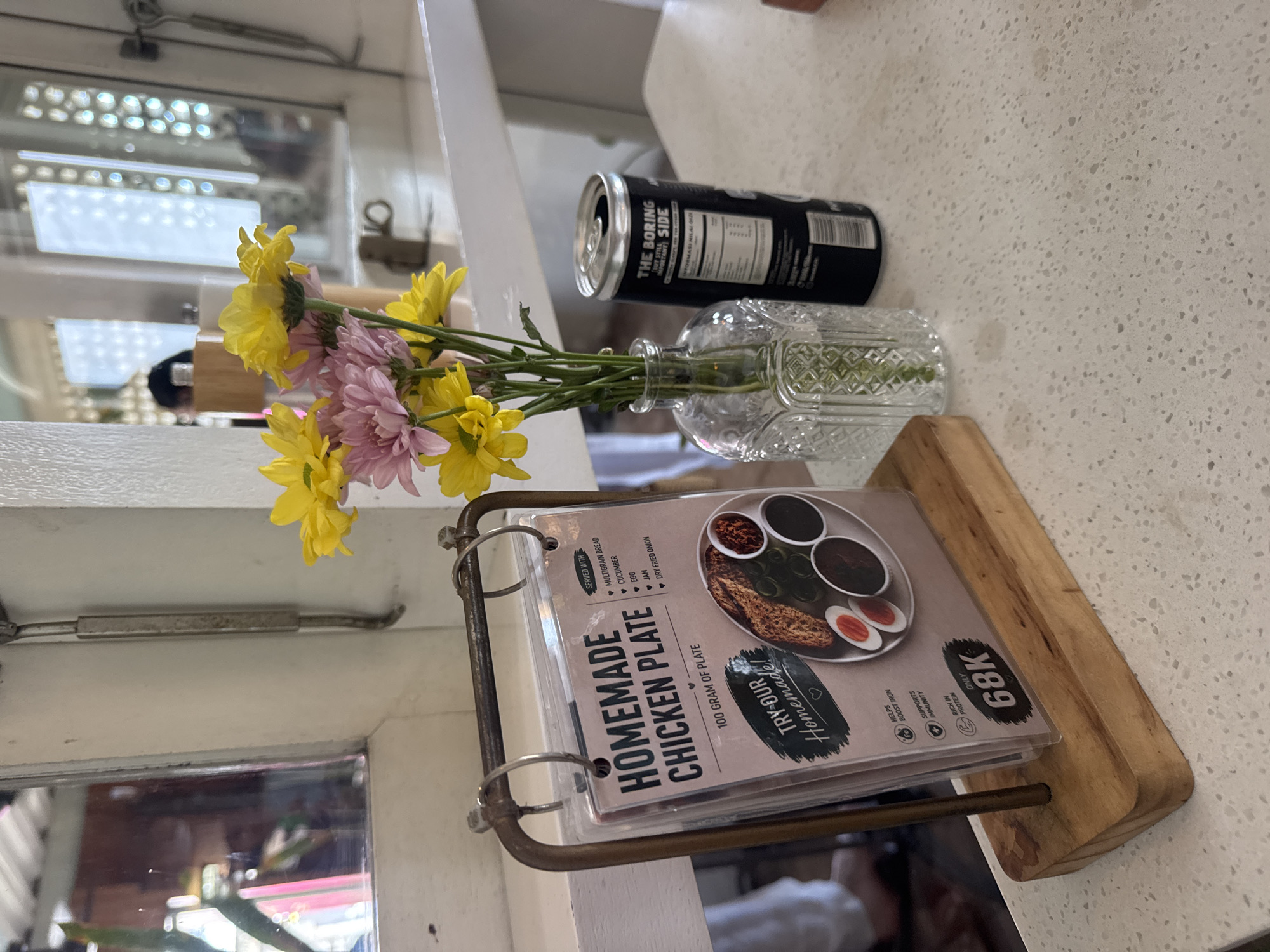 A low-angle medium shot captures a table setting at a cafe, likely in Kuta Selatan, Indonesia. In the foreground, a wooden stand holds a menu displaying a "HOMEMADE CHICKEN PLATE." The menu, protected by plastic, lists its contents as "MULTIGRAIN BREAD, CUCUMBER, EGG, JAM, DRY FRIED ONION." Below this, the price "68K" is prominently displayed. To the right of the menu, a glass vase holds a bouquet of yellow and pink chrysanthemums. Adjacent to the vase is a black can of "THE BORING SIDE" beverage, with its top open. The table surface is a speckled light-colored countertop. In the background, a white wall and a mirror are visible, reflecting a window with an intricate pattern and a person in silhouette. The overall lighting suggests an indoor daytime setting.