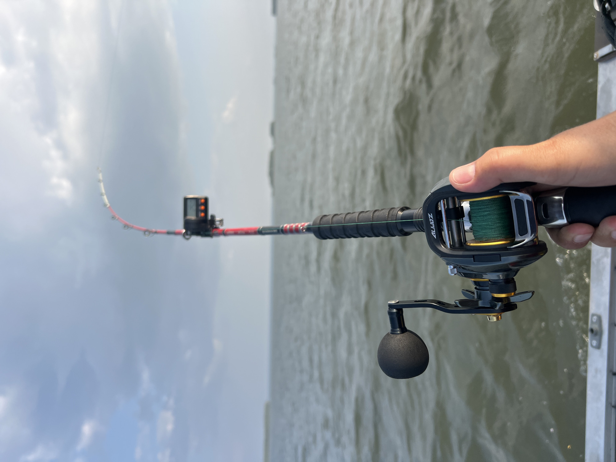A close-up, first-person perspective shot features a hand holding a fishing rod over the water. The fishing rod is black and red with a digital counter attached. The reel is black and gold with green fishing line. The background is a body of water with a cloudy sky. The location is not specified to be in the city of Novopichugovo, Russia, as this information is not present in the image.