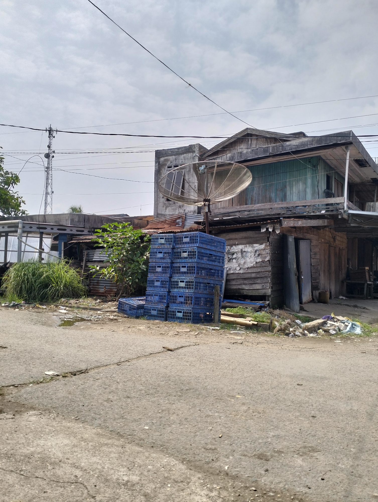 An exterior shot captures a weathered, multi-story wooden building in Serdang Jaya, Indonesia, against a cloudy sky. The building, with its peeling paint and corrugated metal roof, appears aged and somewhat dilapidated. In front of the building, a stack of blue plastic crates is piled high, and a satellite dish is mounted on the side. Tall, dry grass grows to the left of the structure, and the ground in front of the building is a dirt road with scattered debris, including puddles of water and piles of trash. A utility pole with numerous wires is visible in the background, and other structures and vegetation are present, adding to the rustic and somewhat unkempt appearance of the scene.