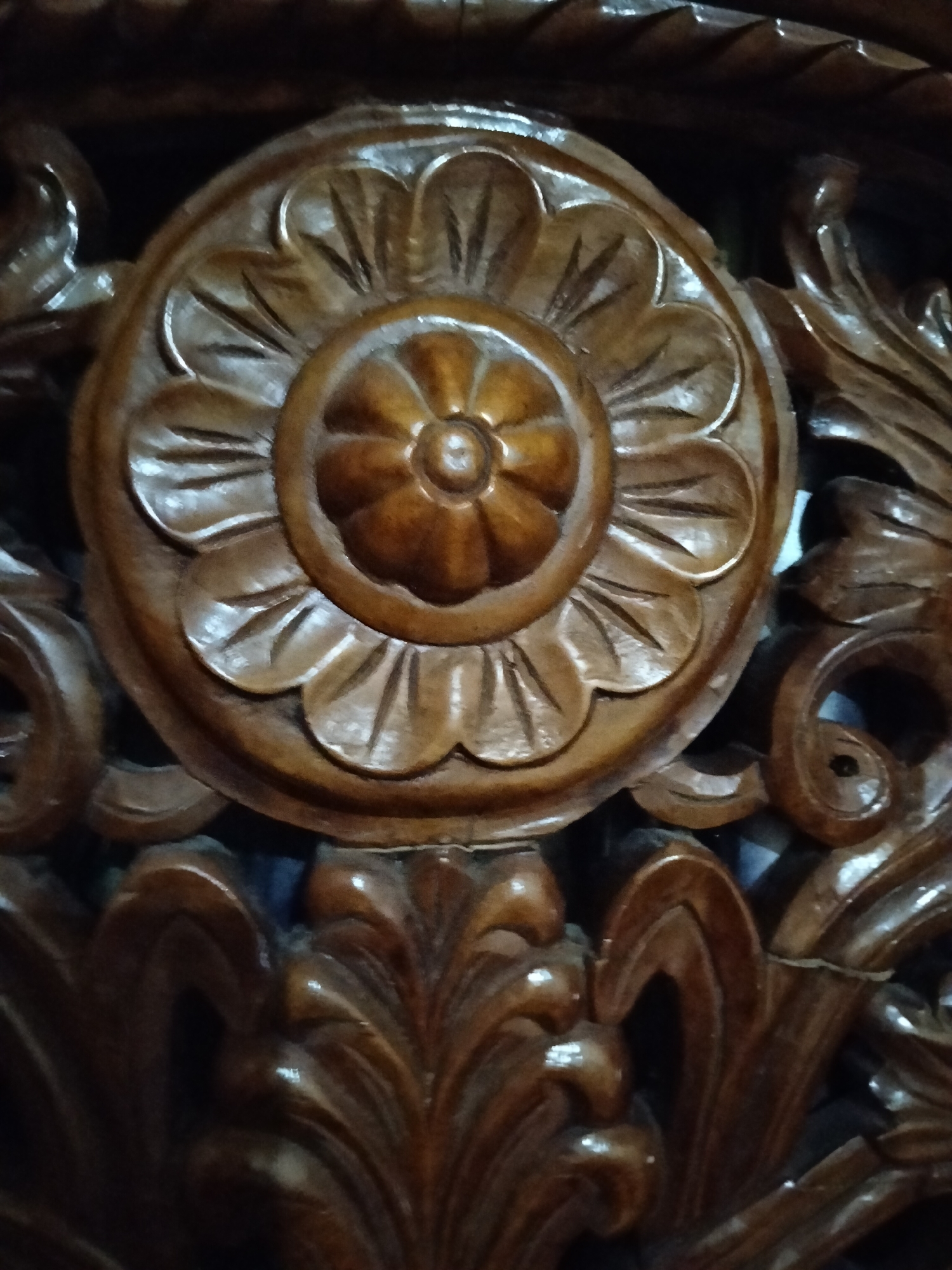 The image shows a close-up of dark brown, intricately carved wooden furniture. The central focus is a circular motif resembling a flower with multiple petals and a smaller circular center, all rendered in wood. Surrounding this central design are flowing, leaf-like carvings and swirling decorative elements. The wood appears to be polished, reflecting light in places, highlighting the depth and detail of the carvings. The background is dark and out of focus, suggesting the furniture is likely indoors, possibly in a dimly lit room. There are no people or discernible actions captured in the image. The image is not geotagged and does not provide information about its location within the city of Payagaboh, Indonesia.