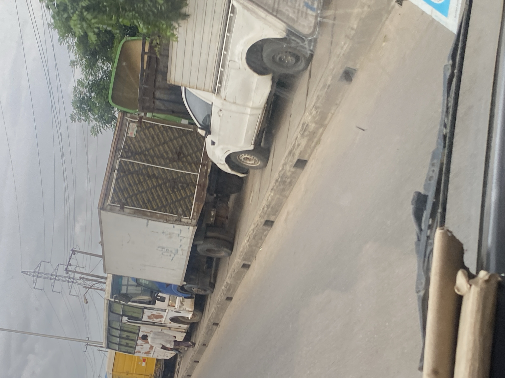 A group of vehicles, including a yellow truck, a white bus, a blue truck with a white cargo box, a green truck, and a white pickup truck, are lined up on a street in Lagos, Nigeria. The street appears to be a major thoroughfare with electrical pylons and overhead wires visible in the background. The vehicles are parked along a concrete curb with a gutter. A person in a white shirt is walking near the white bus. The sky is overcast. The scene suggests a traffic or parking situation in an urban environment.