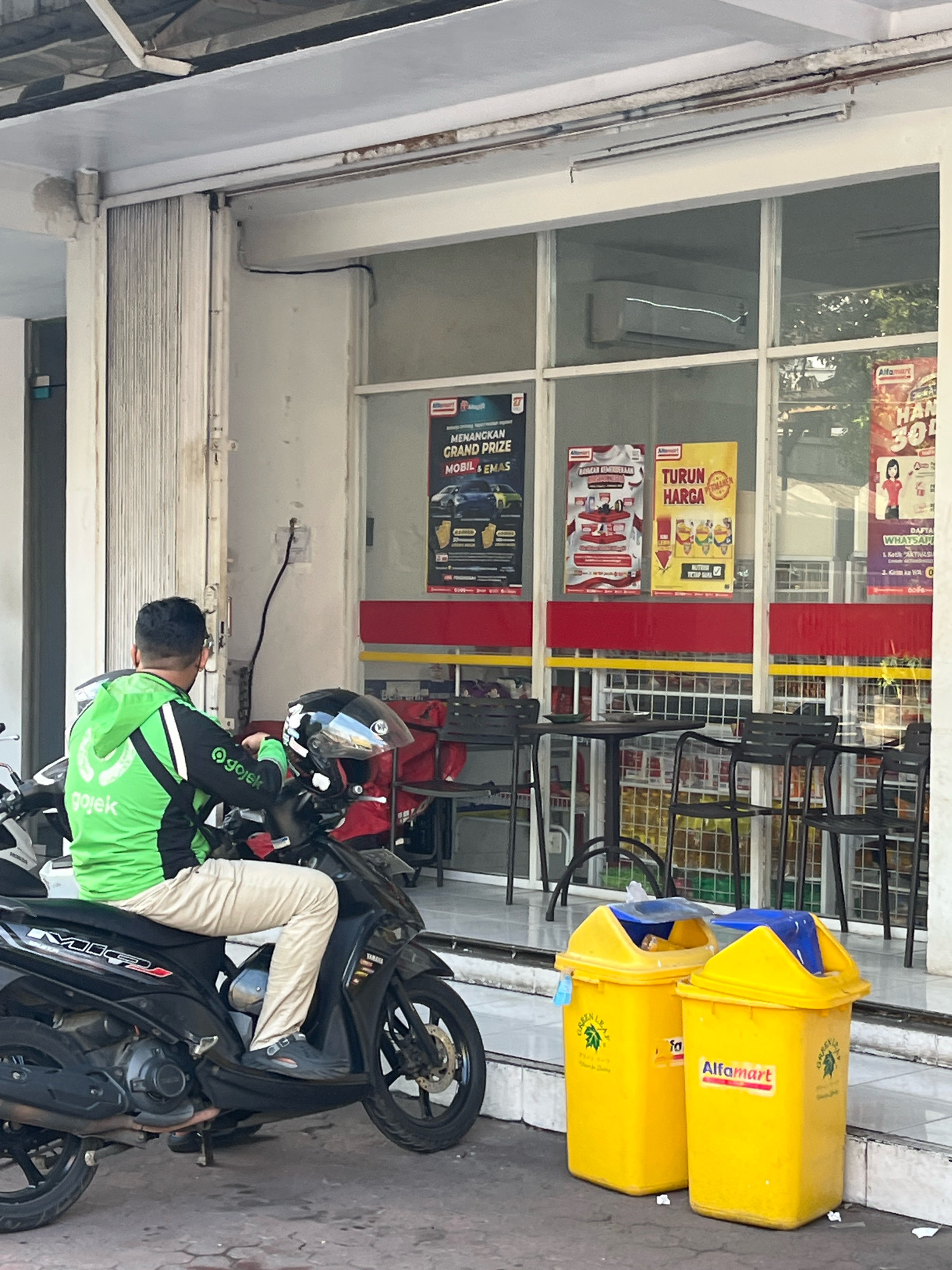 In Yogyakarta, Indonesia, a person wearing a bright green Gojek jacket and light-colored pants sits on a black motorcycle parked in front of a minimarket. The Gojek rider has a black helmet on the seat beside them. The minimarket's storefront features large windows displaying various promotional posters and products. Two bright yellow trash bins with blue lids are placed outside the store. The ground is paved with cobblestones, and a red and yellow striped awning extends over the storefront. The scene appears to be an everyday street view in a commercial area.