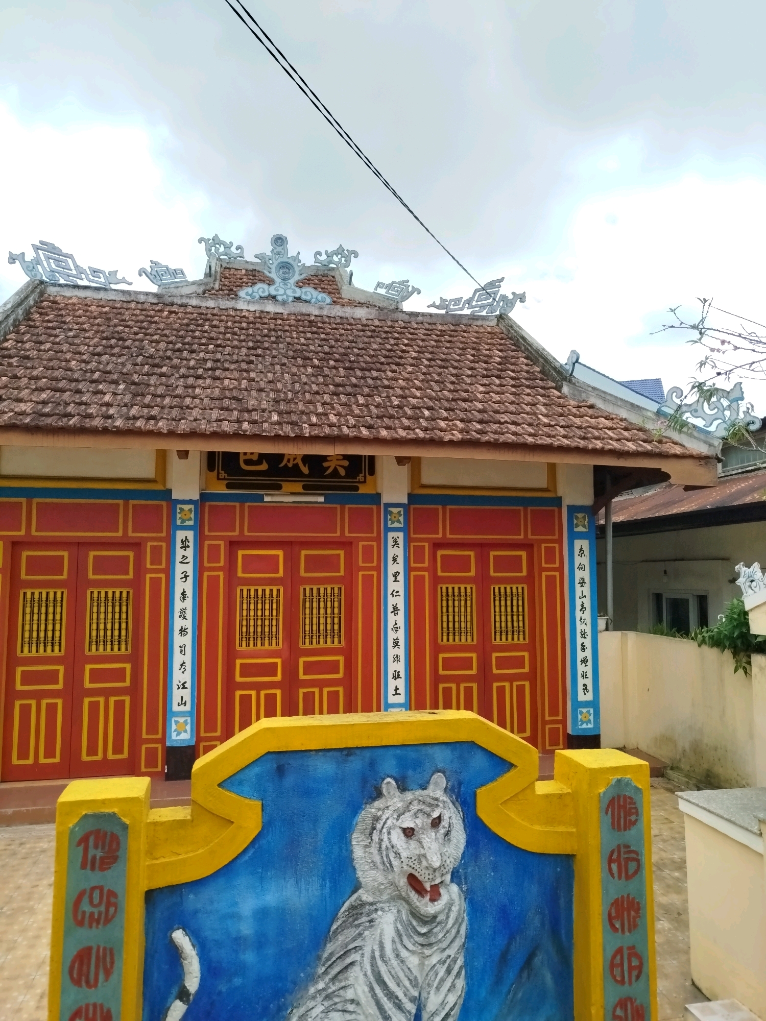 The image shows the facade of a building, likely a temple or shrine, with a traditional East Asian architectural style. The building features a red tiled roof with ornate white decorative elements along the eaves. The walls of the building are painted in vibrant colors, with red double doors adorned with yellow geometric patterns and grilles. Vertical blue panels with white Chinese characters flank the doors. In the foreground, a yellow and blue low wall displays a relief of a white tiger with red eyes and an open mouth, painted against a blue background. The ground appears to be paved with light-colored tiles. A power line is visible stretching across the sky, which is overcast with clouds. The scene is set outdoors, with a hint of surrounding vegetation and other buildings in the background. The presence of Chinese characters suggests a cultural connection to Chinese or Vietnamese traditions. The style of architecture, the decorative elements on the roof, and the relief of the tiger are indicative of a temple or shrine, commonly found in Vietnamese cities like Da Lat. The overall atmosphere is one of cultural and religious significance.