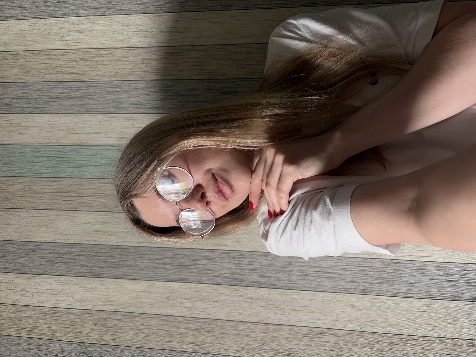 A woman with blonde hair is lying on a wooden floor. She is wearing glasses and a white t-shirt. Her nails are painted red. The floor is made of planks with varying shades of gray and brown. There is no indication of the city of Chaikovsky, Russia.