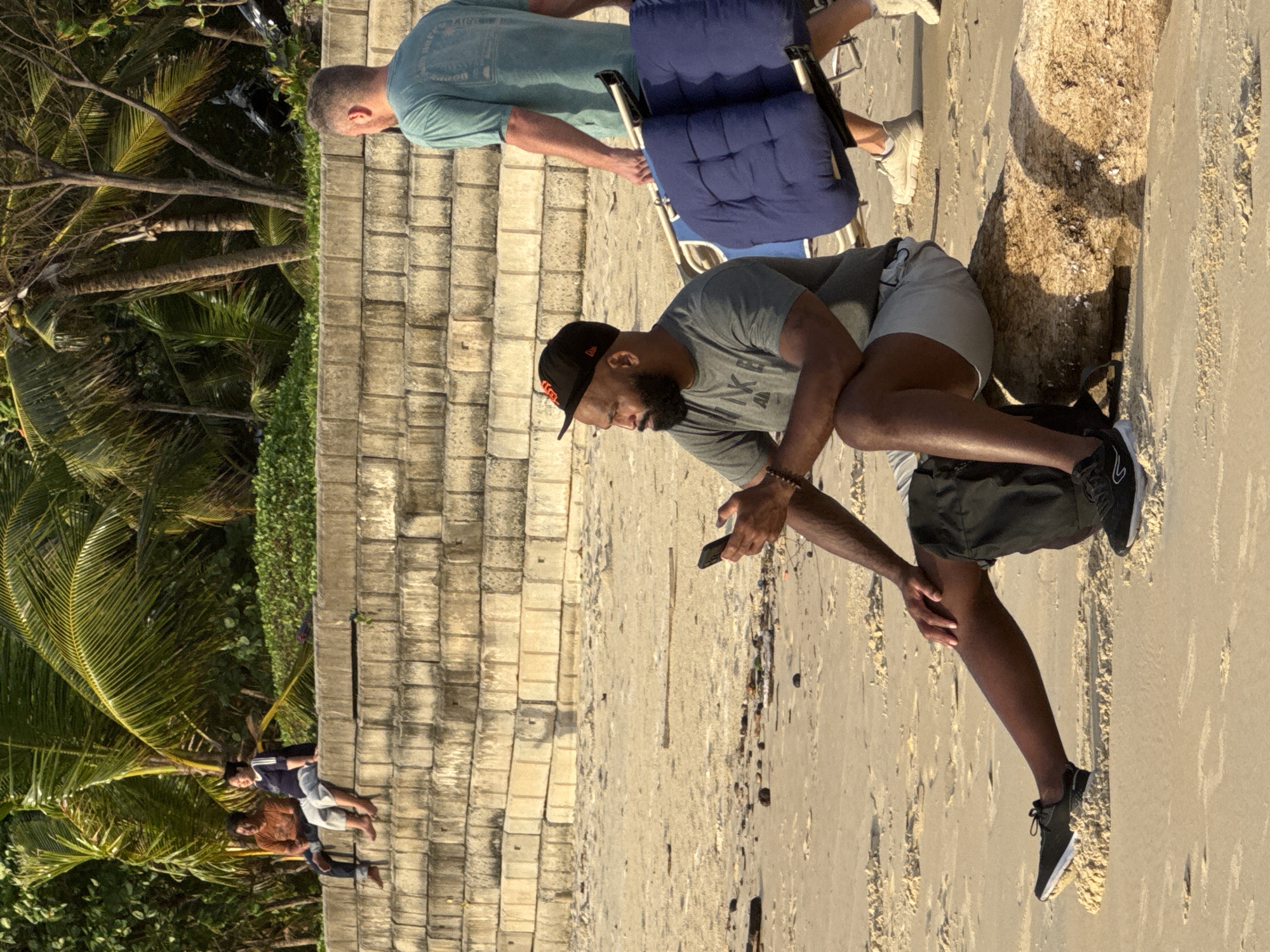 In Karon, Thailand, a medium shot captures a beach scene with a seawall in the background and palm trees overhead. In the foreground, a man with a beard and a black baseball cap is sitting on a log on the sand, looking at his phone. He is wearing a gray t-shirt and light-colored shorts. To the right, another man in a teal t-shirt is walking away from the camera, carrying a folded blue chair with a metal frame. In the background, two people are sitting on the seawall. The sand is scattered with small pieces of debris. The overall lighting suggests it is late afternoon.