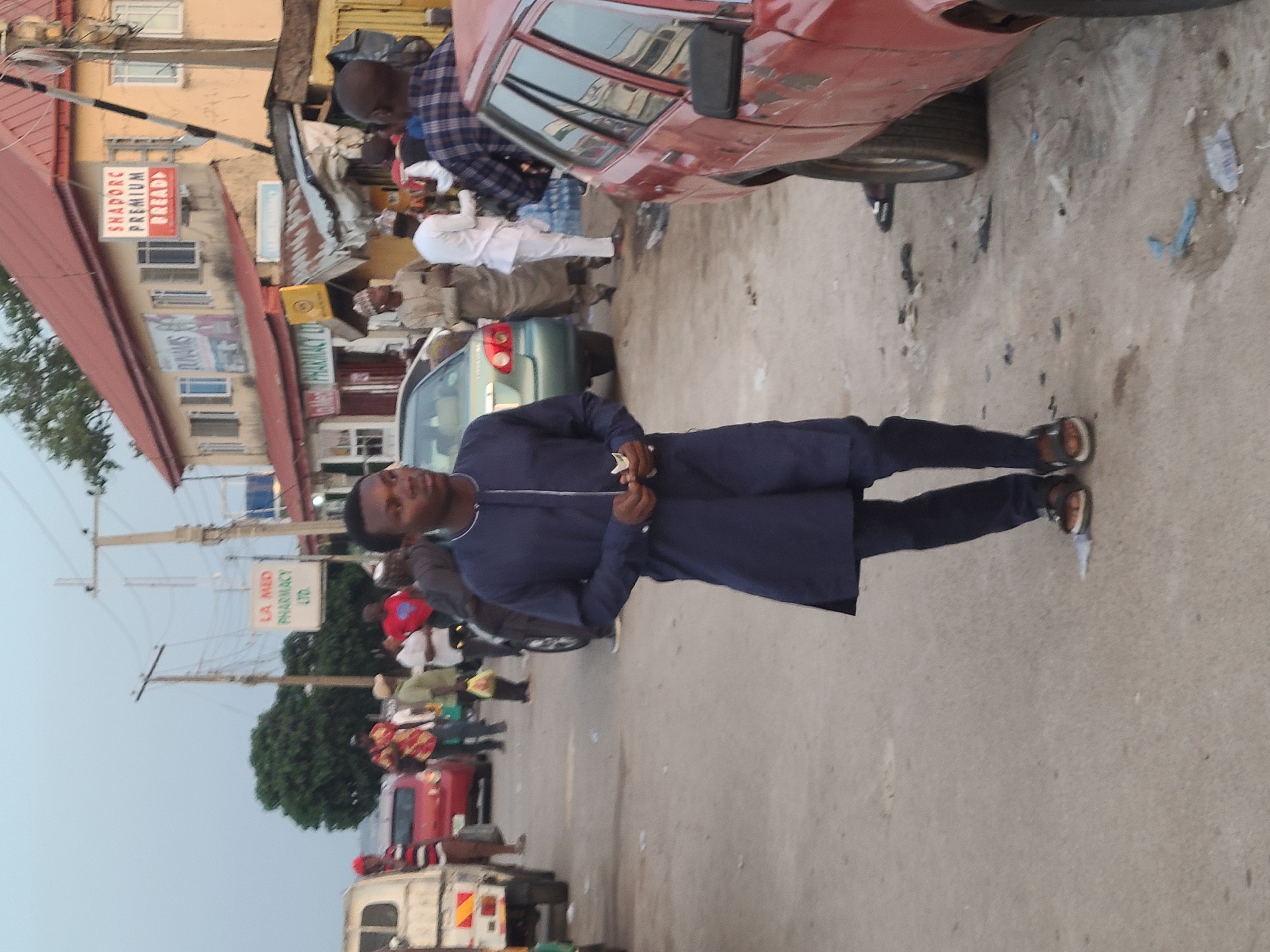 A street scene in Unknown, Unknown. A man in a blue outfit stands in the foreground. Cars and pedestrians are in the background. Buildings and street signs are visible.