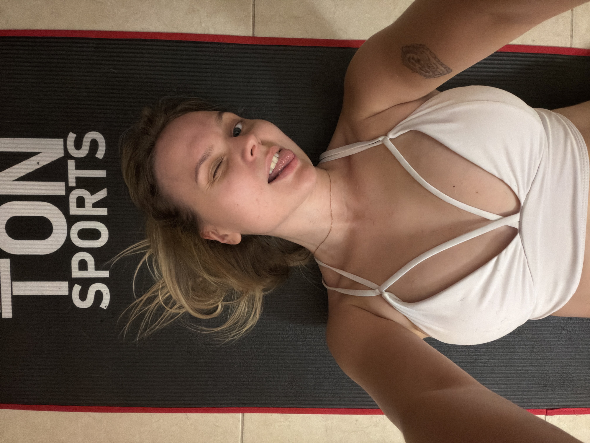 A young woman is lying on a black and red exercise mat with the words "TON SPORTS" printed on it. She is wearing a white sports bra and sticking her tongue out while winking. She has a tattoo on her left upper arm. The background shows a tiled floor. The image does not appear to have been taken in Tel-Aviv, Israel.
