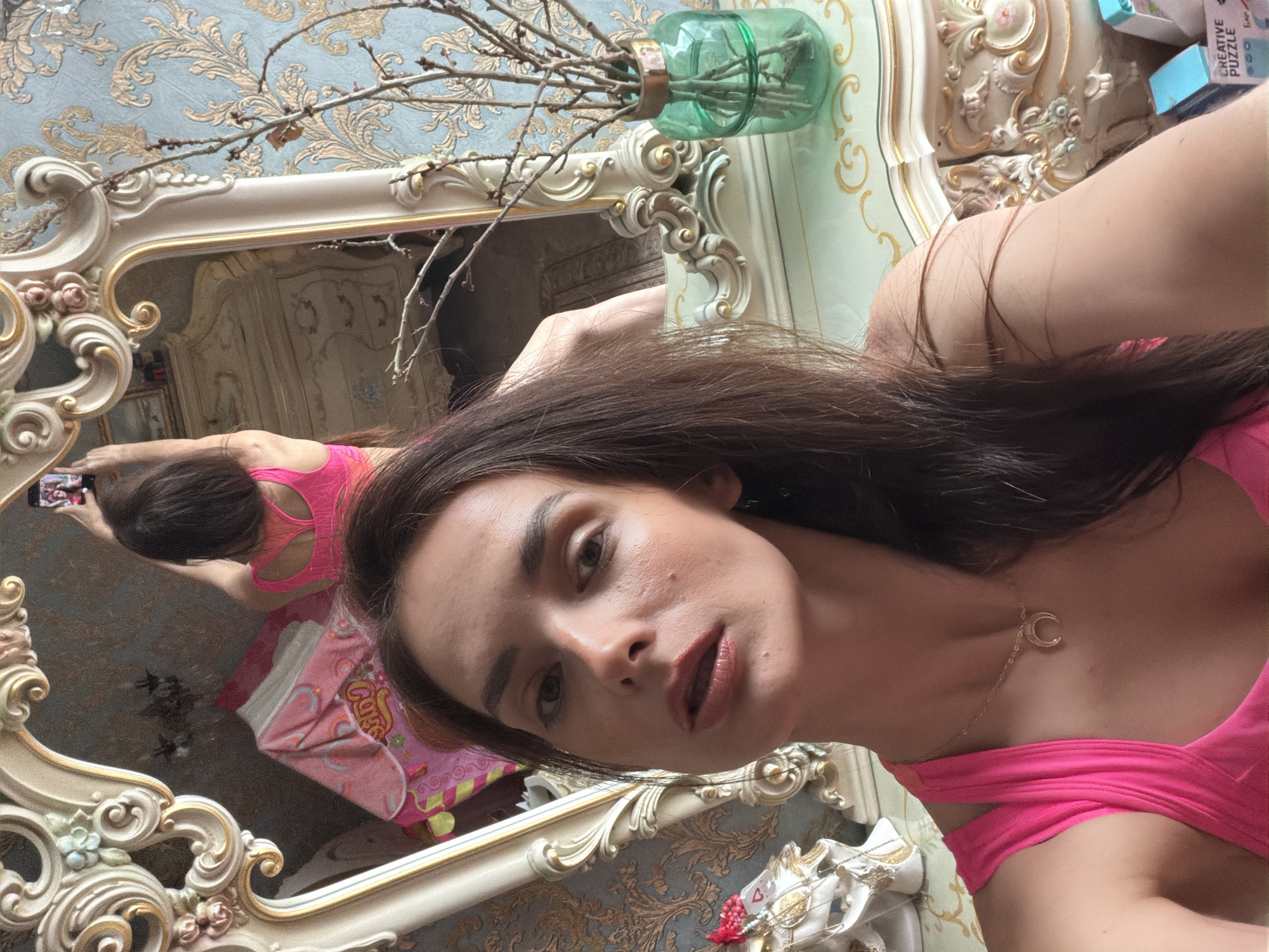 A woman with long, dark hair, wearing a bright pink tank top and a crescent moon necklace, takes a selfie in a mirror. She is in a room with ornate, antique-style furniture. The mirror reflects the back of her head and shoulders, as well as a pink blanket draped over a piece of furniture. To the right of the mirror, a green glass vase holds bare branches. The wall behind the furniture has a textured, damask-like wallpaper in blue and gold. A small part of a box labeled "Creative Puzzle" is visible in the lower right corner.