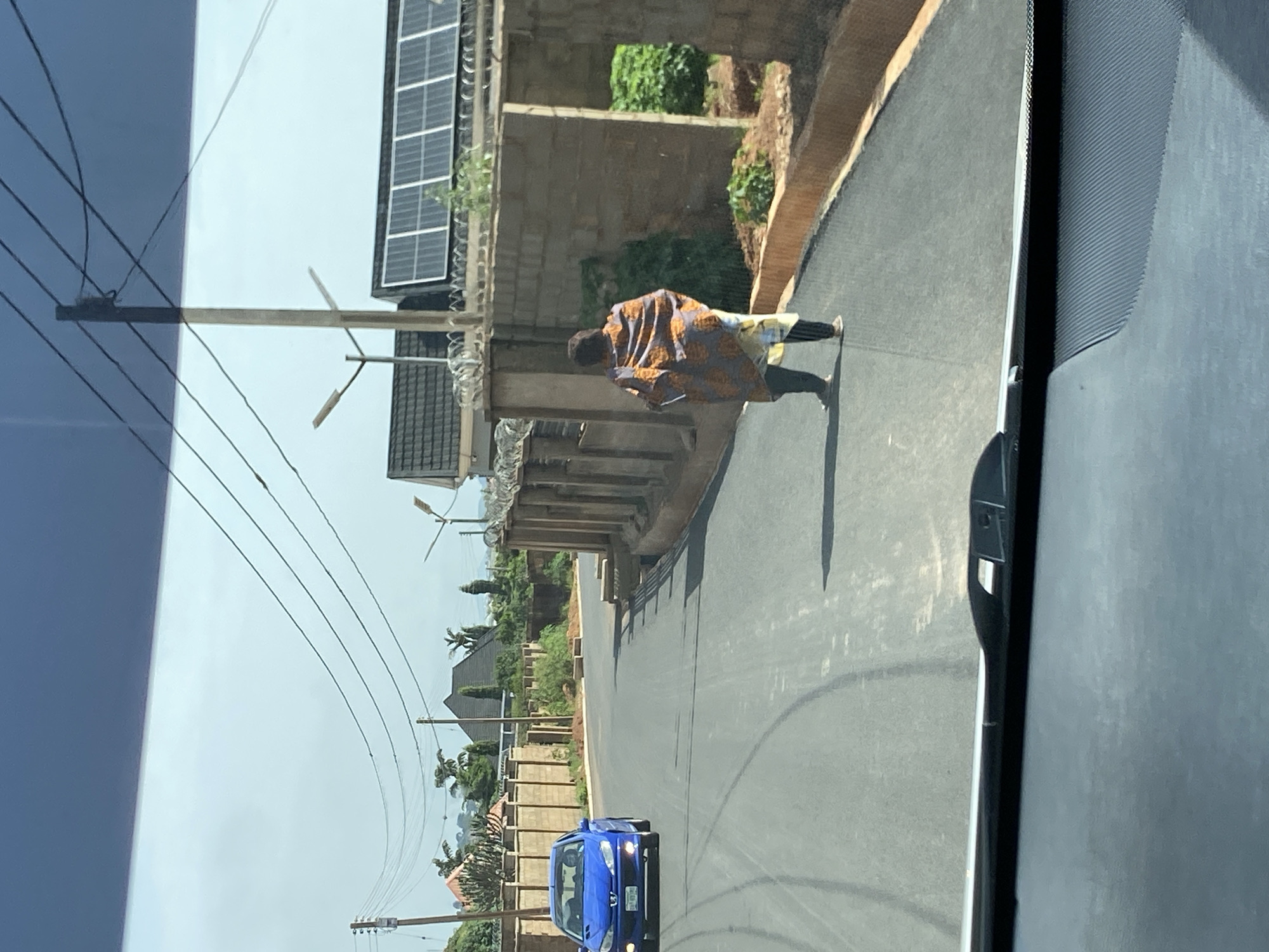 A blue Peugeot 206 hatchback is parked on the left side of a newly paved asphalt road in Bar Jirgi Summa, Nigeria. The car faces forward, with its headlights on. The driver is not visible. A woman is walking away from the camera on the right side of the road, wearing a colorful patterned wrapper and dark pants. She is walking towards a brick wall with a metal fence on top, which borders a property with solar panels on the roof of a building. The road is lined with concrete barriers and some sparse vegetation. In the background, there are trees and houses, with overhead power lines crisscrossing the sky. The sky is overcast.