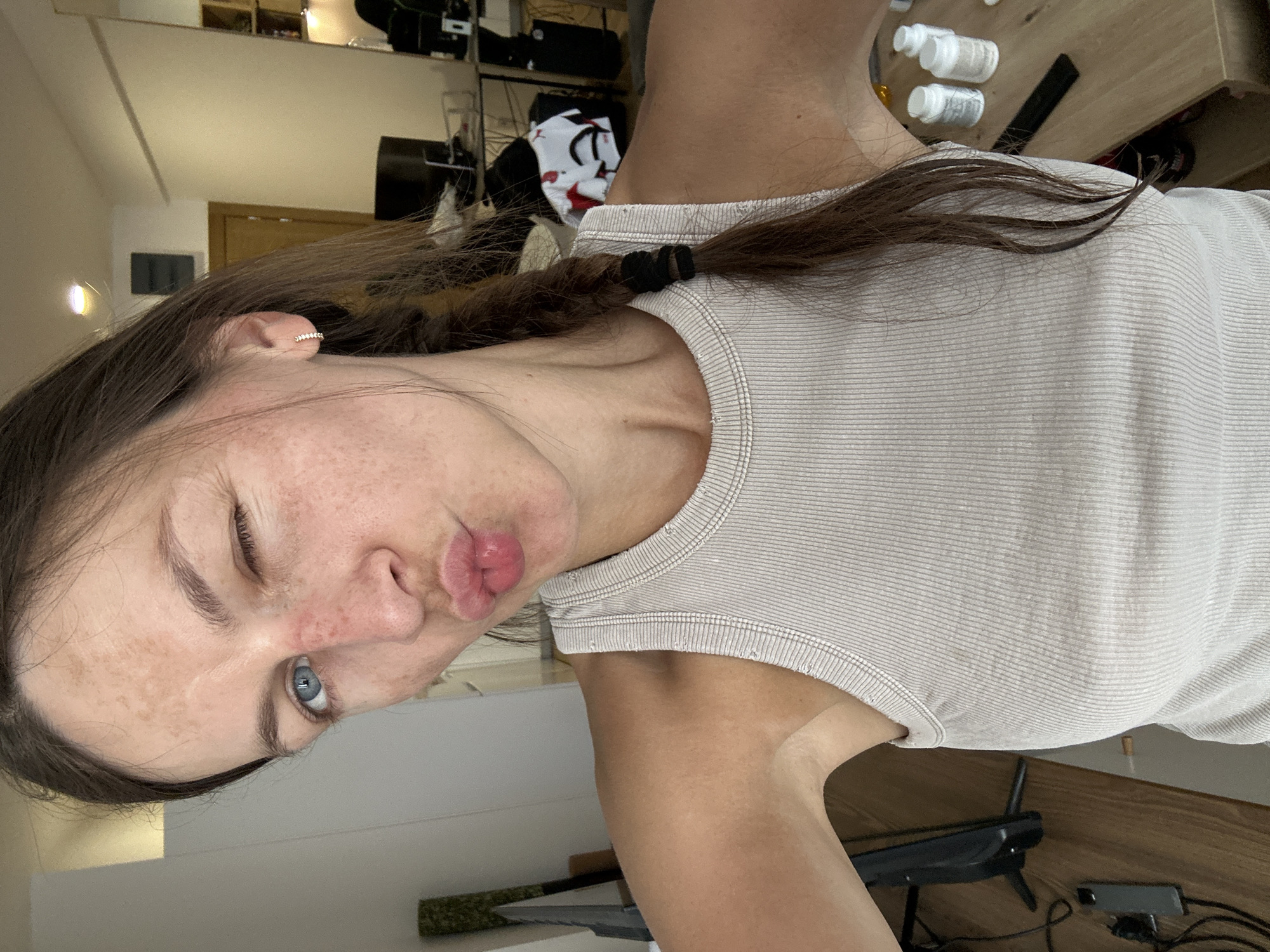 The image shows a selfie of a woman with dark brown hair in a braid and blue eyes. She is wearing a light-colored ribbed tank top. She is winking and puckering her lips. The background appears to be an indoor setting with furniture and objects that suggest a living space. There is no indication of the location being Donji Pobori, Montenegro.