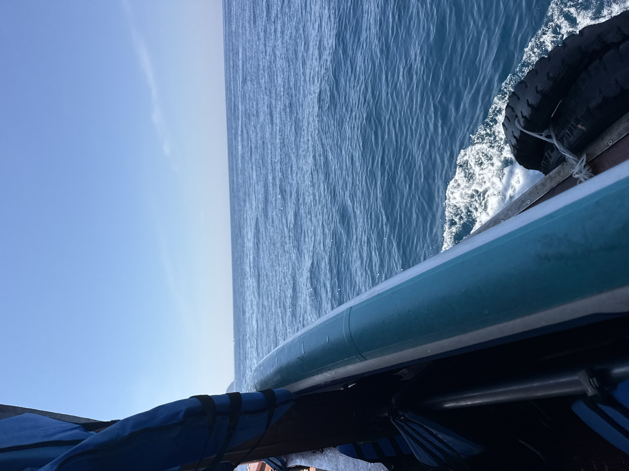 This image shows a view from the side of a boat moving through the ocean. The boat has a blue and white railing. On the left side of the image, there are blue life vests. The ocean is a deep blue and there is a clear sky above. The boat appears to be traveling along the coast of Nha Trang, Vietnam, given the typical scenery of boats and the ocean in that region. The water is calm with small waves. There are no people visible in the frame.