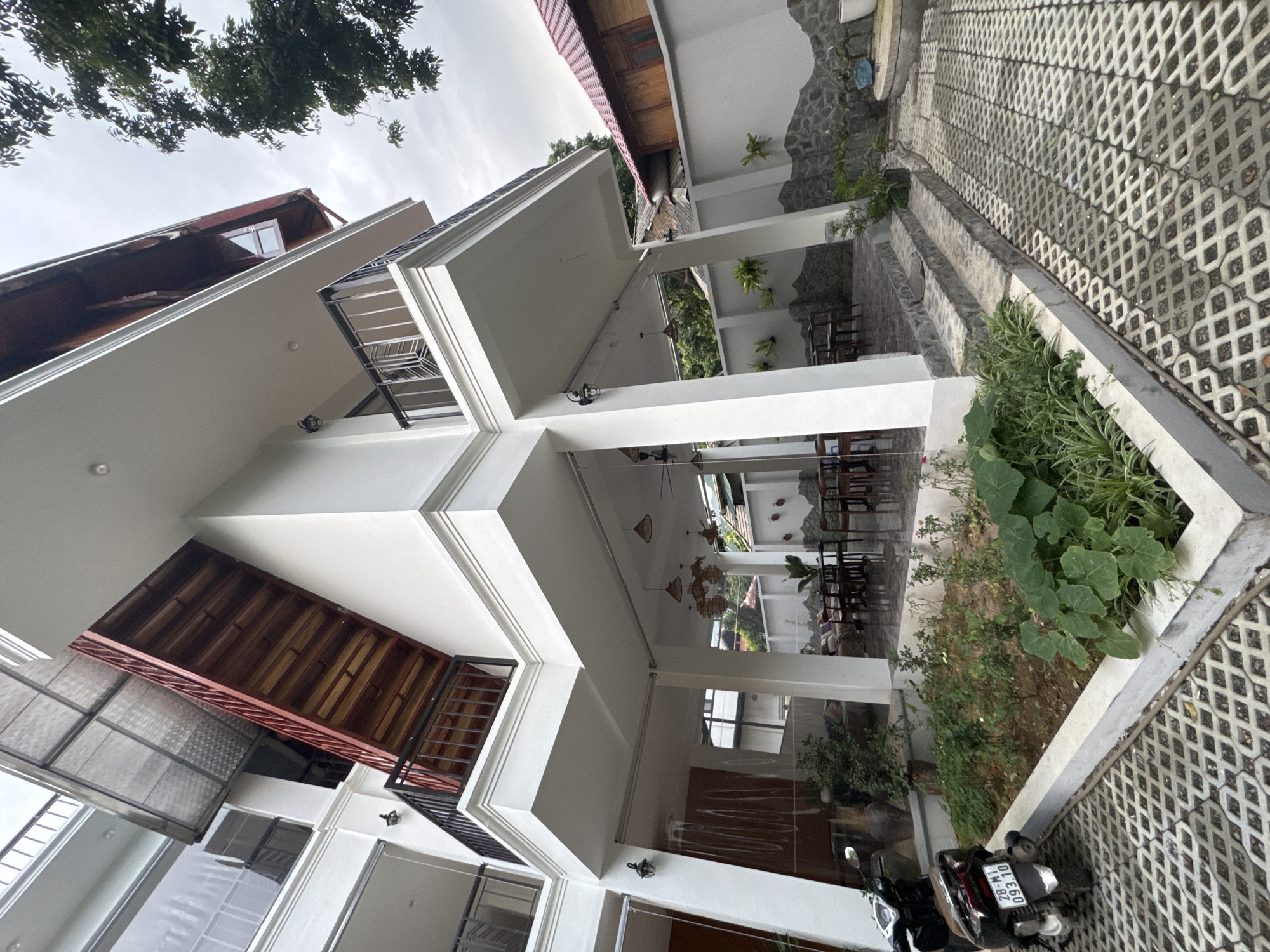 This image depicts the exterior of a multi-story building, possibly a guesthouse or a small hotel, with a focus on its architectural design and outdoor spaces. The building has white painted walls and several balconies with black railings. The ground is paved with a distinctive interlocking stone pattern.
In the foreground, a portion of a parked motorcycle is visible, along with its license plate which reads "28-N1 093.10". Adjacent to the paving is a raised planter bed filled with lush green vegetation, including large-leafed plants and smaller flowering plants. The setting appears to be outdoors, with trees visible in the upper left corner and in the background. The architecture suggests a tropical or subtropical climate.
Further into the scene, we can see outdoor seating areas with wooden tables and chairs, indicating a space for guests to relax or dine. Hanging light fixtures, possibly lanterns, are visible under covered walkways or balconies. The overall atmosphere is one of a tranquil, possibly resort-like environment. The sky is overcast, suggesting a cloudy day. The image is taken from a low angle, looking upwards and to the side, giving a dynamic perspective of the building. No people are clearly visible in the image. The location context provided is Mai Chau, Vietnam.