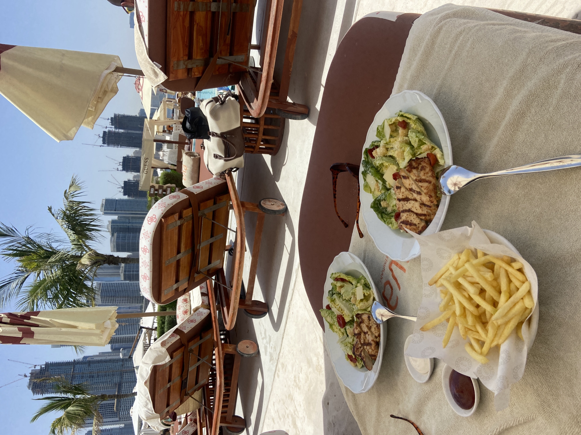 The image shows a lunch setting in Dubai, United Arab Emirates. Two plates are visible, one with a chicken salad and the other with fries, along with small containers of ketchup and mayonnaise. The food is placed on a light-colored towel atop a brown surface. Eyeglasses lie nearby. In the background, there are beach chairs, umbrellas, palm trees, and numerous high-rise buildings and skyscrapers under construction, a common feature of Dubai's urban landscape. The sky is clear. The scene is well-lit, suggesting it's daytime.