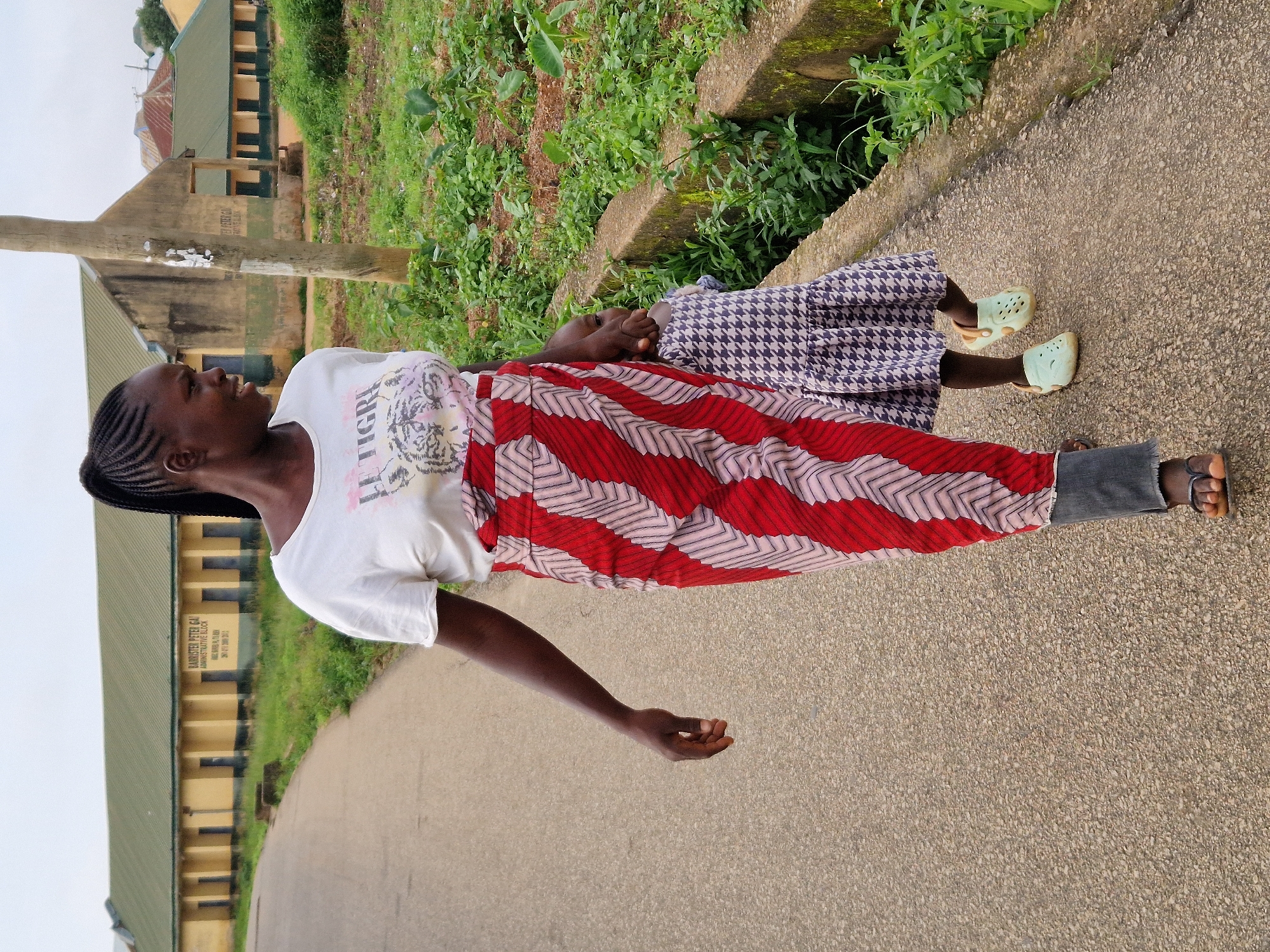 In Kabong, Nigeria, a woman with dark skin and her hair braided into a ponytail walks on a paved road with a young child. The woman wears a white t-shirt with a graphic design and a long, red and pink patterned wrap skirt. She is also wearing dark pants and dark sandals. The child, who is holding the woman's hand, wears a purple and white checkered dress and light blue clogs. They are walking past a concrete drainage ditch and a patch of green vegetation. In the background, a long, single-story building with a green roof and yellow walls stands under an overcast sky. A wooden pole is visible to the right of the woman.
