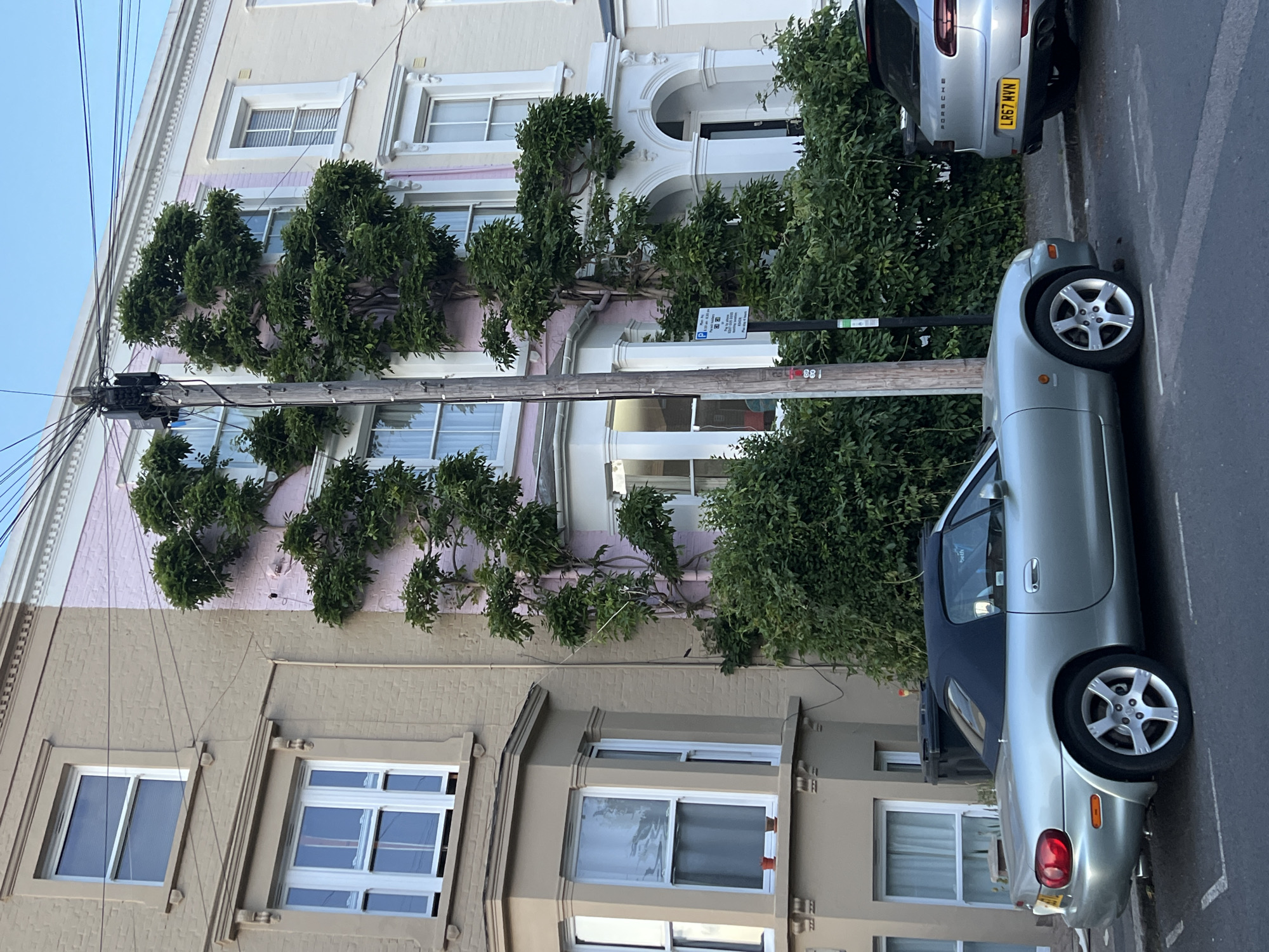 The image shows a street scene in London, United Kingdom. Two cars, a silver Porsche and a silver Mazda MX-5, are parked on the right side of the street. On the left, there are terraced houses with bay windows and decorative facades. The houses are beige and light pink. Trees or vines are growing along the front of the houses, obscuring the windows. A utility pole with power lines is visible on the left side of the frame. A street sign is positioned between the two cars, partially obscured by greenery.