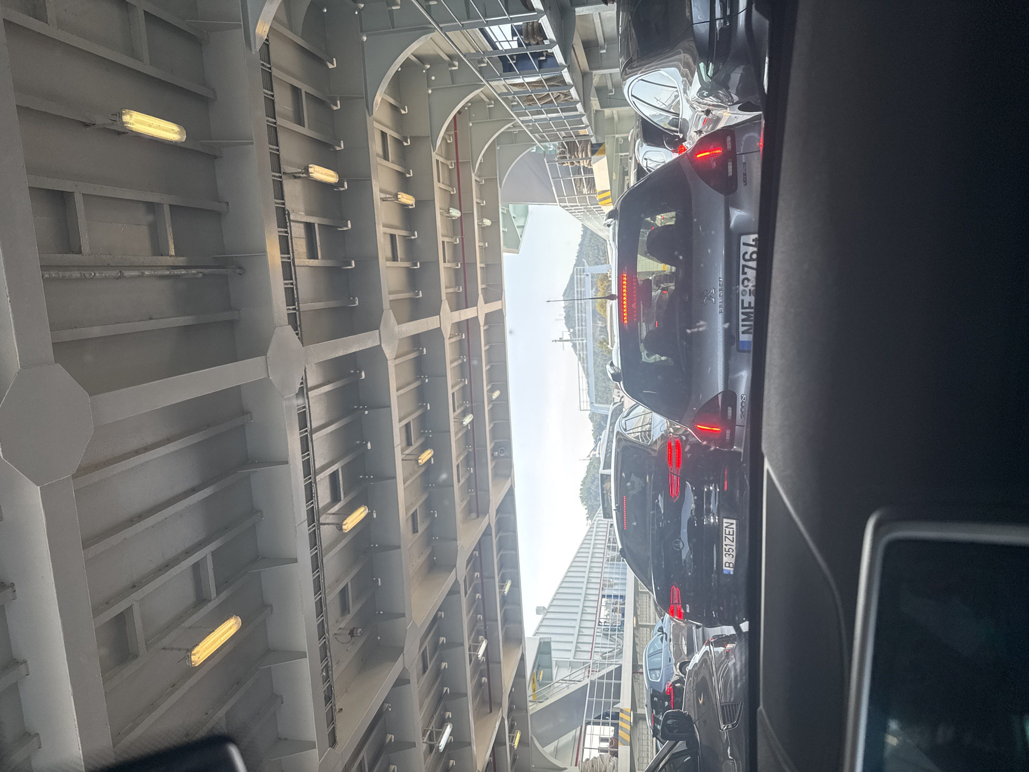 The image depicts a traffic jam on the deck of a ferry, likely in the port of Thassos, Greece, given the destination context provided in the prompt. The view is from inside a vehicle, looking forward through the windshield and out at other cars queuing on the ferry. The ferry's interior is characterized by a metallic ceiling with exposed beams and rows of fluorescent lights. Several cars are visible, including a black Mercedes-Benz with license plate "B 351ZEN" and a silver Peugeot with license plate "NME 8764". The cars are closely packed, indicating a large number of vehicles on board. In the background, glimpses of the port and the island's landscape, including a hill, are visible through an opening. The lighting suggests it is daytime. There are no clearly visible people in the foreground, though silhouettes can be seen within some of the cars.
