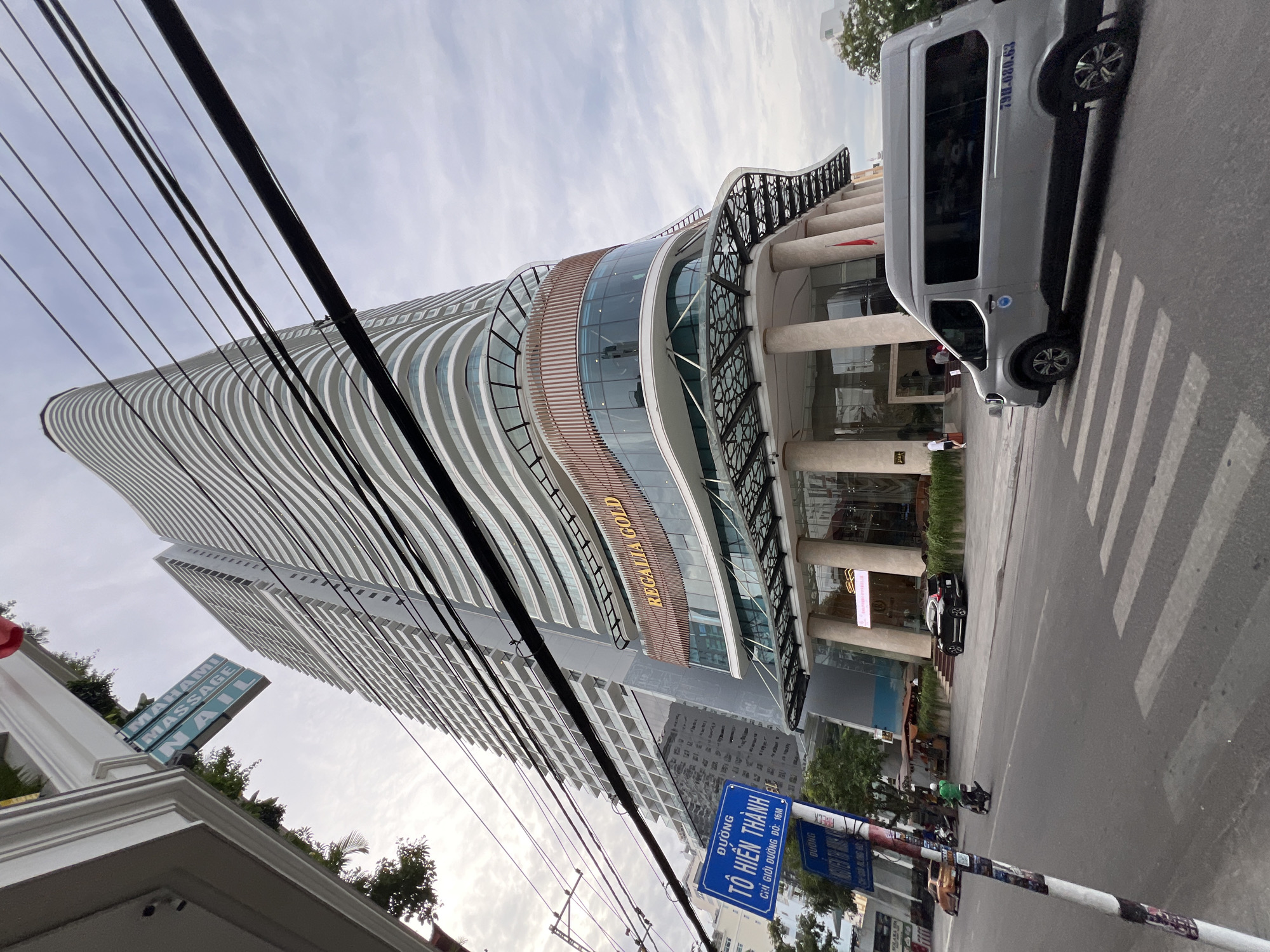 In Nha Trang, Vietnam, a low-angle shot captures a modern, multi-story building with a curved facade and white balconies. The building's base features a glass-enclosed entrance with large pillars and an ornate canopy. Above the entrance, the words "REGALIA GOLD" are visible in gold lettering. In the foreground, a gray van with tinted windows drives across a crosswalk on a paved street. A blue street sign with white Vietnamese text, likely indicating a street name like "TO HIEN THANH," is positioned to the left of the van. Overhead, a tangle of black electrical wires crisscrosses the sky, obscuring the pale, cloudy atmosphere. To the left of the building, a white sign with "MAHAMI MASSAGE NAIL" in blue lettering is attached to a pillar. People are visible entering the building and on a sidewalk. The street also shows a black car parked and a motorcycle with a rider.
