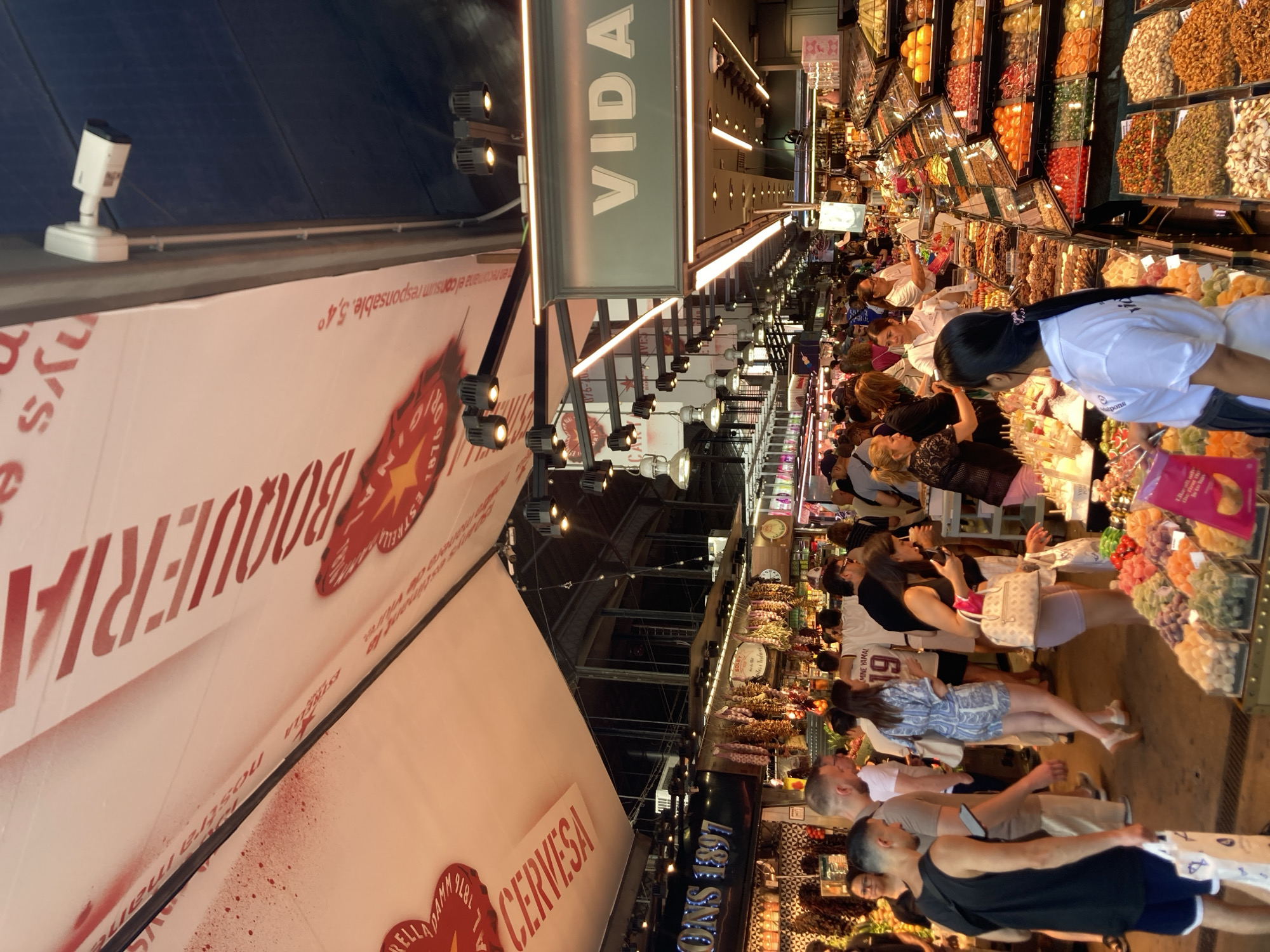 This media file captures a bustling scene inside a large indoor market, likely in Barcelona, Spain, given the text visible. The image is oriented vertically, making it appear as if the viewer is looking down at the market floor from a high vantage point or a tilted angle.
The primary subjects are a crowd of people browsing through stalls filled with a vibrant array of goods. The stalls appear to be selling a variety of items, including dried fruits, nuts, candies, and possibly other food products, showcased in colorful displays. Many individuals are seen looking at the merchandise, interacting with vendors, or walking through the crowded aisles. The overall atmosphere suggests a lively and busy marketplace.
Visible text includes "CERVESA" (Spanish for beer), "BOUQUERIA" which likely refers to La Boqueria Market in Barcelona, and "VIDA" (Spanish for life). There are also signs with numbers like "19" and "1897," and what appears to be a logo with a star. The lighting suggests it might be daytime, with artificial lights illuminating the interior. The people's attire is casual, consistent with a busy day of shopping. Some individuals are wearing uniforms, possibly indicating they are vendors or market staff. The image conveys a sense of a vibrant, culturally rich, and busy commercial environment.