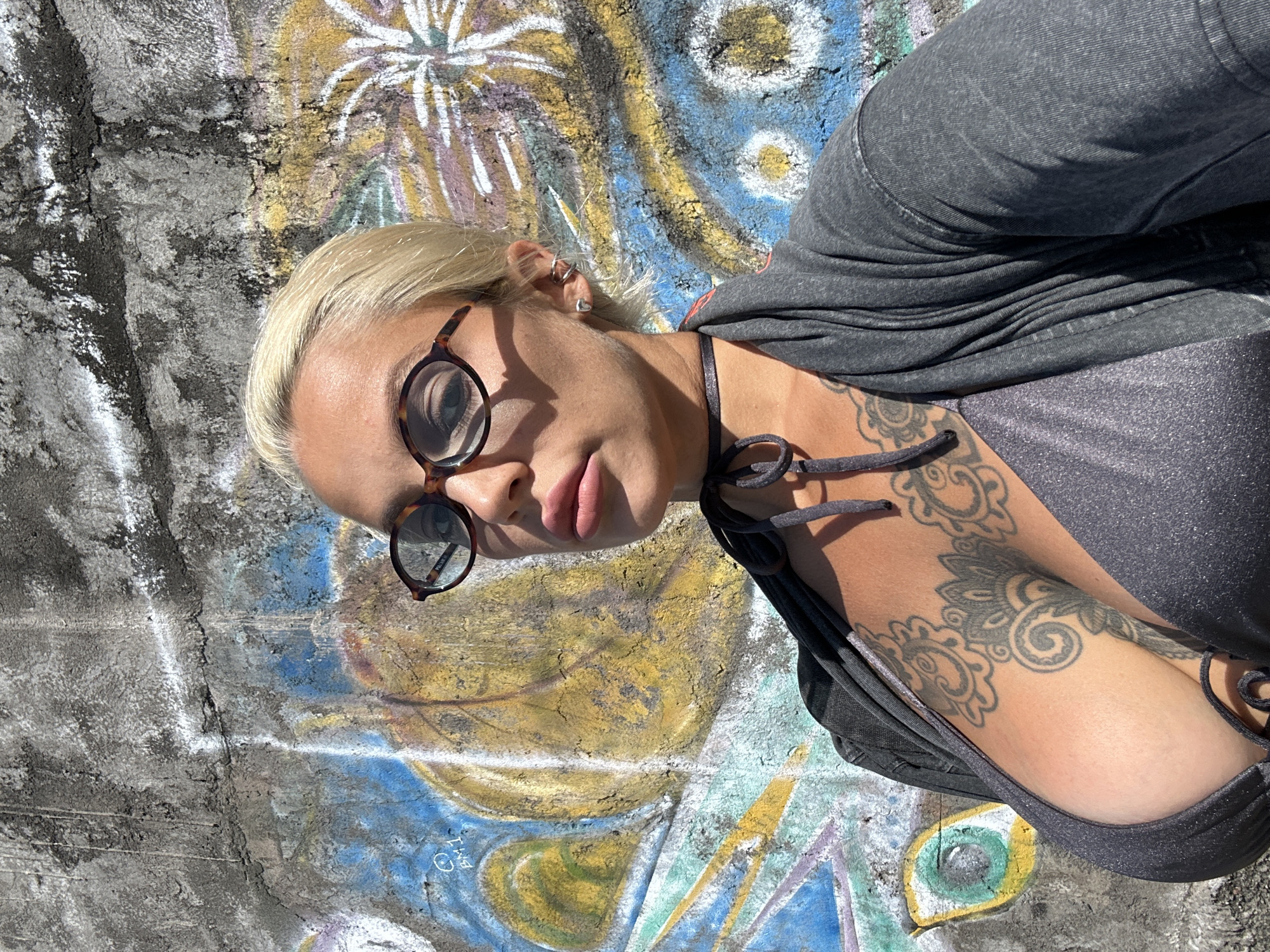 A close-up, low-angle, outdoor photograph features a blonde woman in a grey bikini top and a grey open cardigan, standing in front of a graffiti-covered concrete wall. The woman is wearing tortoise-shell glasses and a dark choker necklace with ties. She has prominent tattoos on her chest and upper torso. Sunlight casts shadows on her face and body. The graffiti on the wall is colorful, with abstract shapes and patterns, including what appears to be an eye. The scene suggests a casual, possibly urban or artistic setting in Kuta Selatan, Indonesia, given the distinct graffiti style often found in such locations.
