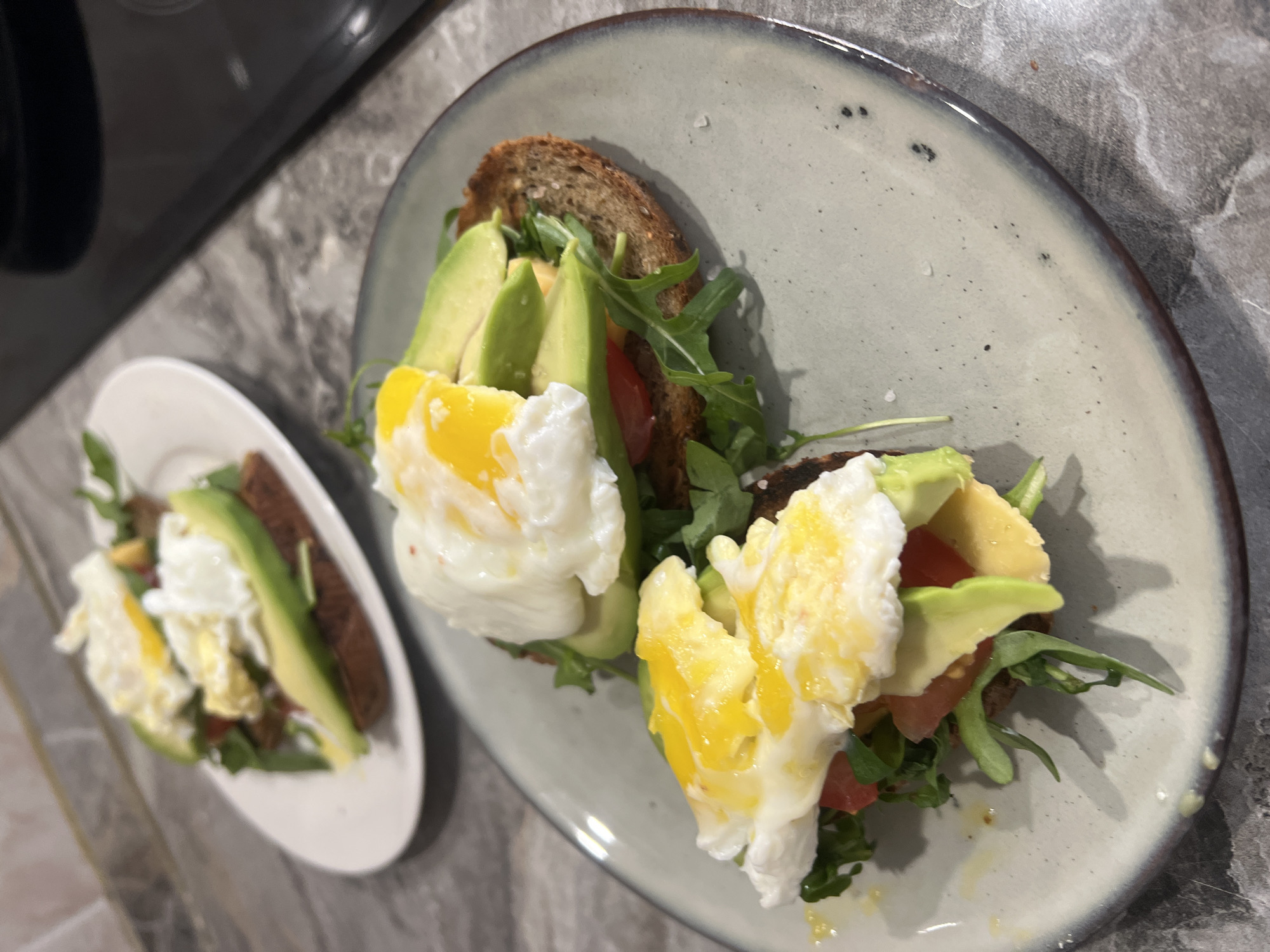 Two plates containing avocado toast with poached eggs are set on a counter. The counter surface has a marble-like pattern. The larger plate, which is grey with a dark rim, holds two slices of toast topped with arugula, sliced tomatoes, avocado slices, and poached eggs with runny yolks. A smaller, white plate contains a similar toast arrangement, though it appears to have only one slice. Part of a black appliance is visible in the upper left corner. The scene is indoors, likely in a kitchen or dining area. The exact location within Khimki, Russia, cannot be determined from the image alone.