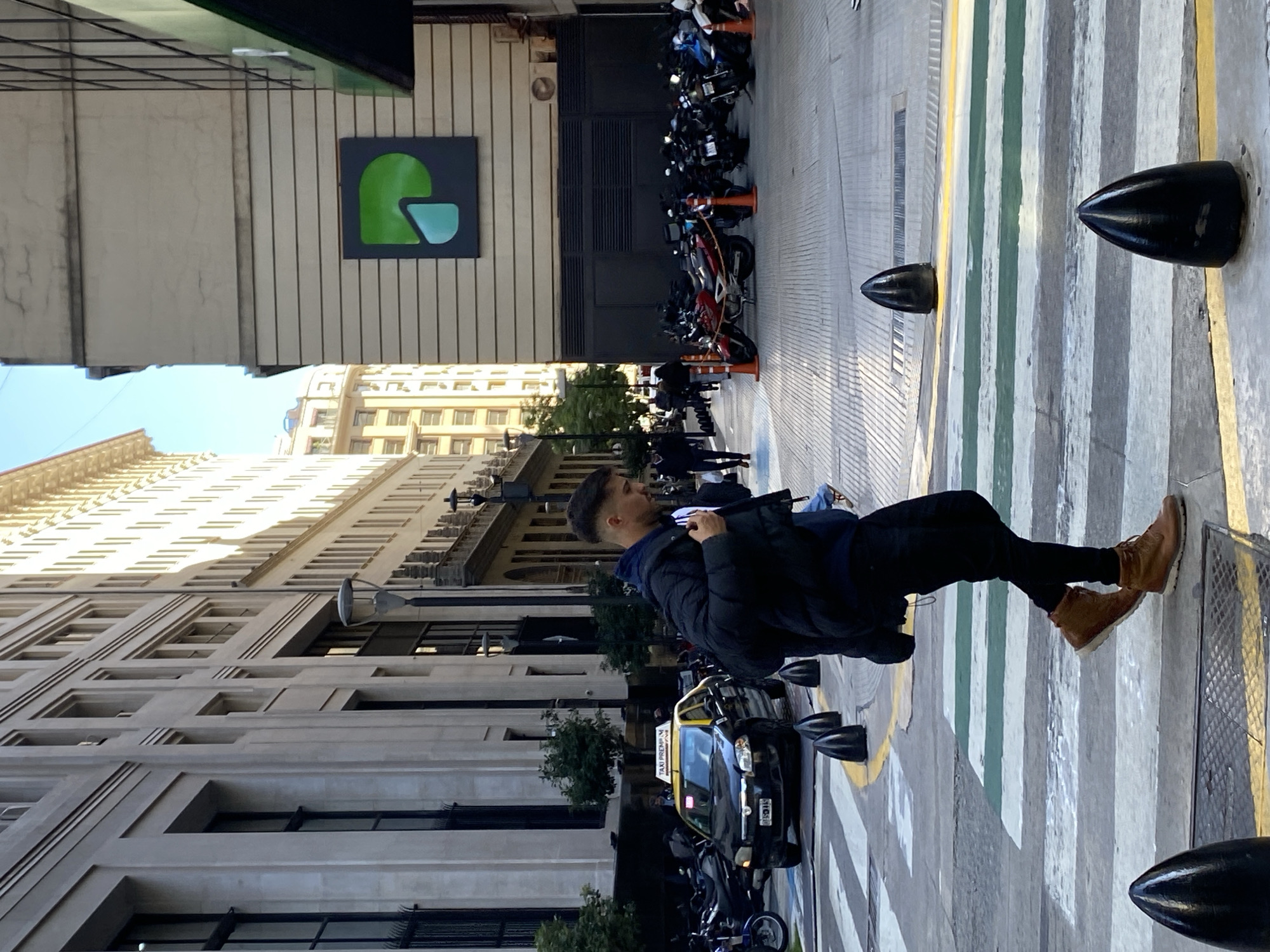 A sunlit urban street scene depicts a young man walking on the right side of the frame, alongside a street and sidewalk. He is dressed in a dark puffer jacket, a blue hoodie, black pants, and light brown lace-up boots, with short, dark hair.
To the left of the man, a black and yellow taxi cab is visible, featuring a "TAXI PREMIU" sign on its roof and a partially legible license plate displaying "BAJ 538". Numerous motorcycles are parked closely together along the street. The street surface is marked with a distinctive pedestrian crossing pattern of alternating grey, green, and yellow stripes. Several black, conical bollards are spaced along the edge of the pavement, separating the street from the sidewalk.
Tall, multi-story buildings line both sides of the street, suggesting a bustling city environment. One building on the left exhibits an older, ornate architectural style. Another building, further down the street, features a noticeable logo: a stylized green shape, possibly resembling an abstract 'P' or a helmet profile with a light blue inner section, set against a dark grey square panel.
The scene is brightly illuminated by natural sunlight, indicating it is daytime on a clear, sunny day, with distinct shadows cast across the pavement and buildings.