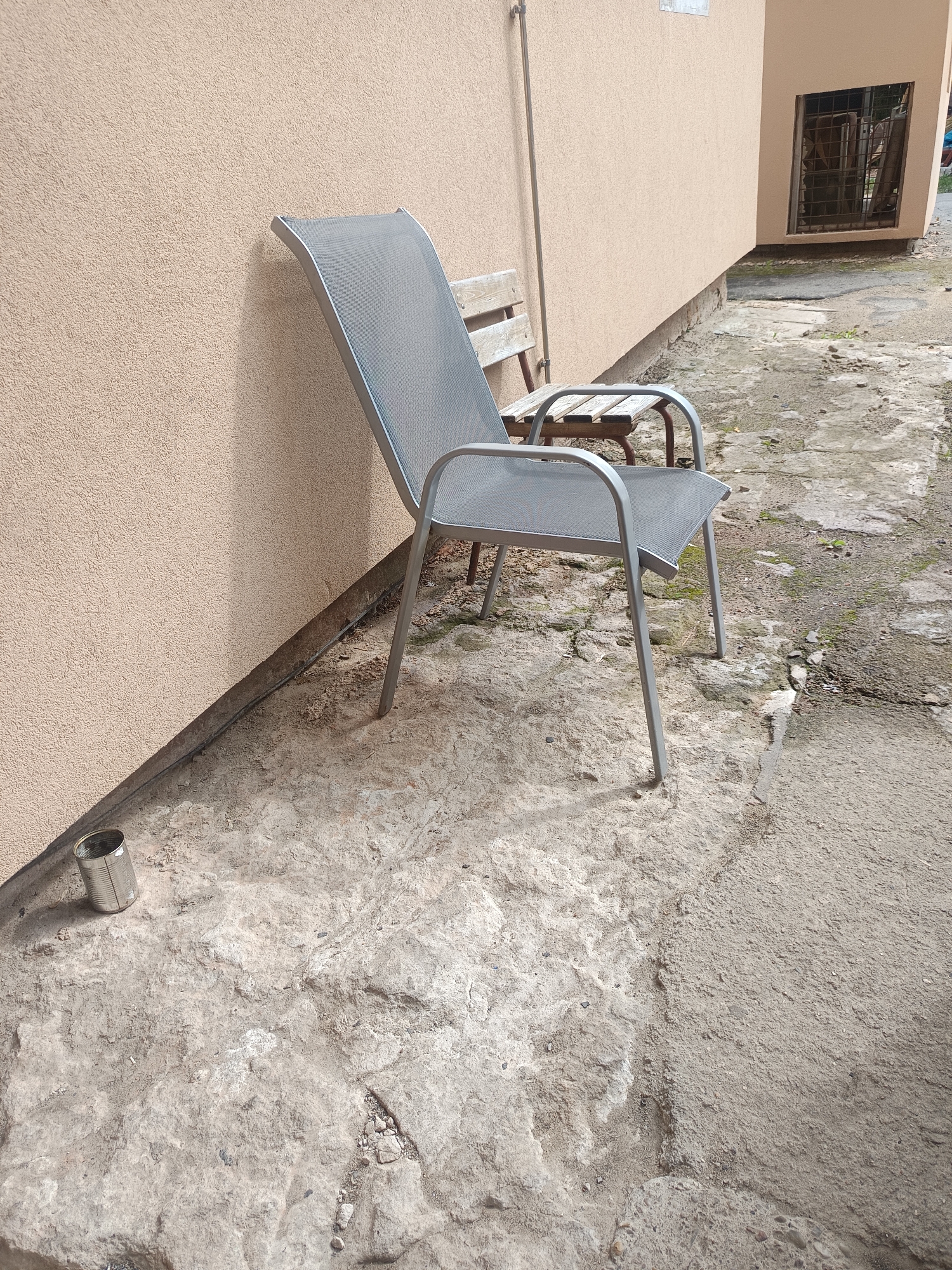 An outdoor scene shows two chairs and a metal can on a rough concrete surface next to a building with a beige stucco wall. One chair, a gray metal and mesh lounger, is positioned at an angle to the wall, its legs resting on the concrete. Behind it is a smaller, rustic wooden bench with metal legs. A dented silver metal can is on the concrete to the left of the lounger. The ground is uneven, with patches of dirt and debris. The building has a window with bars in the upper right corner. The scene appears to be in a courtyard or an alleyway.