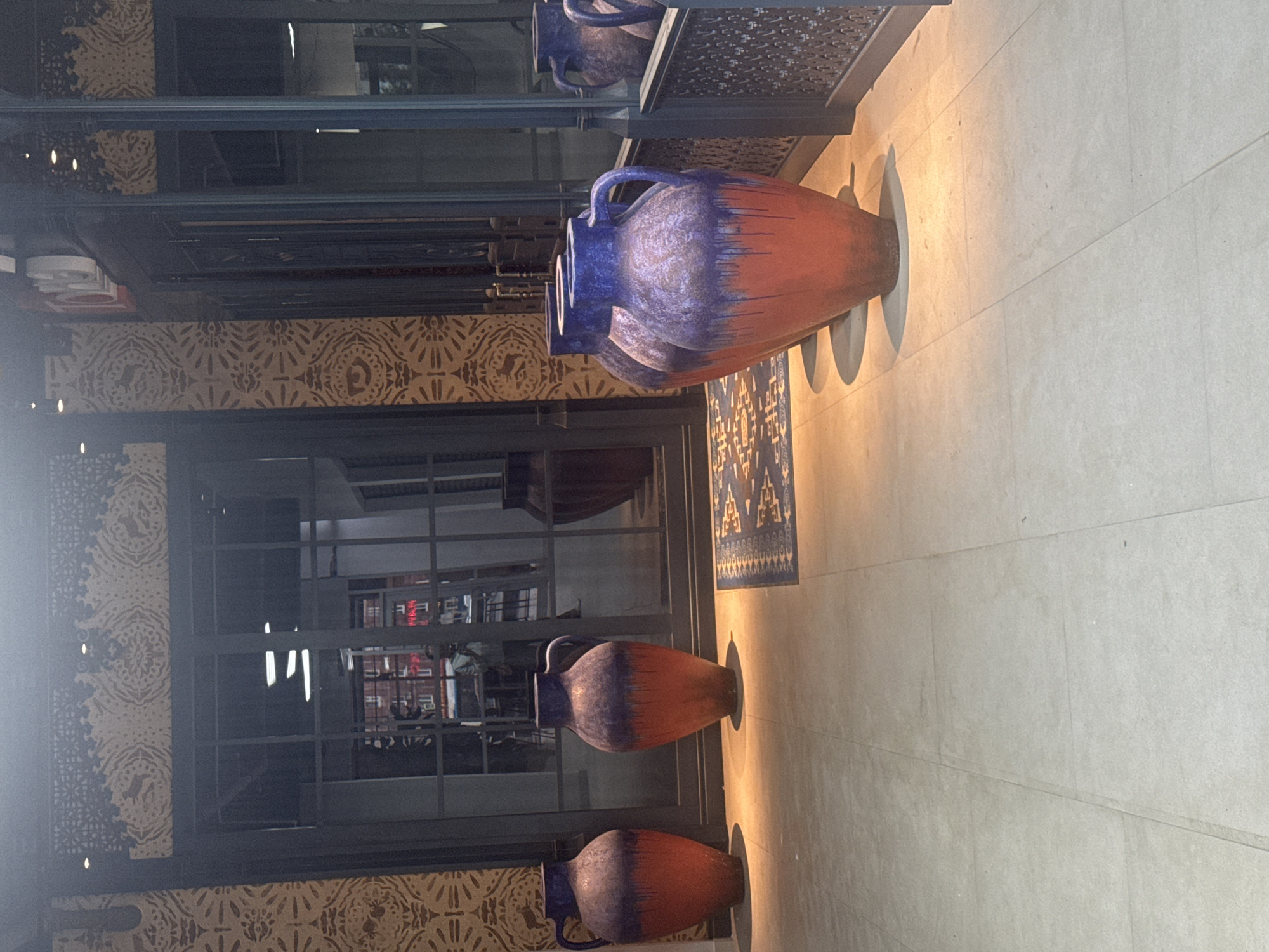 A group of large, decorative ceramic vases are displayed in an entryway. The vases have a gradient color scheme, with a reddish-orange base transitioning to a deep blue at the top, and appear to be glazed with a textured finish. Four vases are prominently visible in the foreground, with two on the left and two more to the right, partially obscured by a reflective surface. Behind the vases, a large glass-paned door allows a view into a dimly lit interior with fluorescent lighting. The floor is tiled with large, light-colored square tiles, and a patterned rug lies in front of the vases. The walls feature decorative stenciled patterns. The scene appears to be an indoor or covered outdoor space, possibly a shop or restaurant entrance. There is no specific indication of the location being Ufa, Russia.