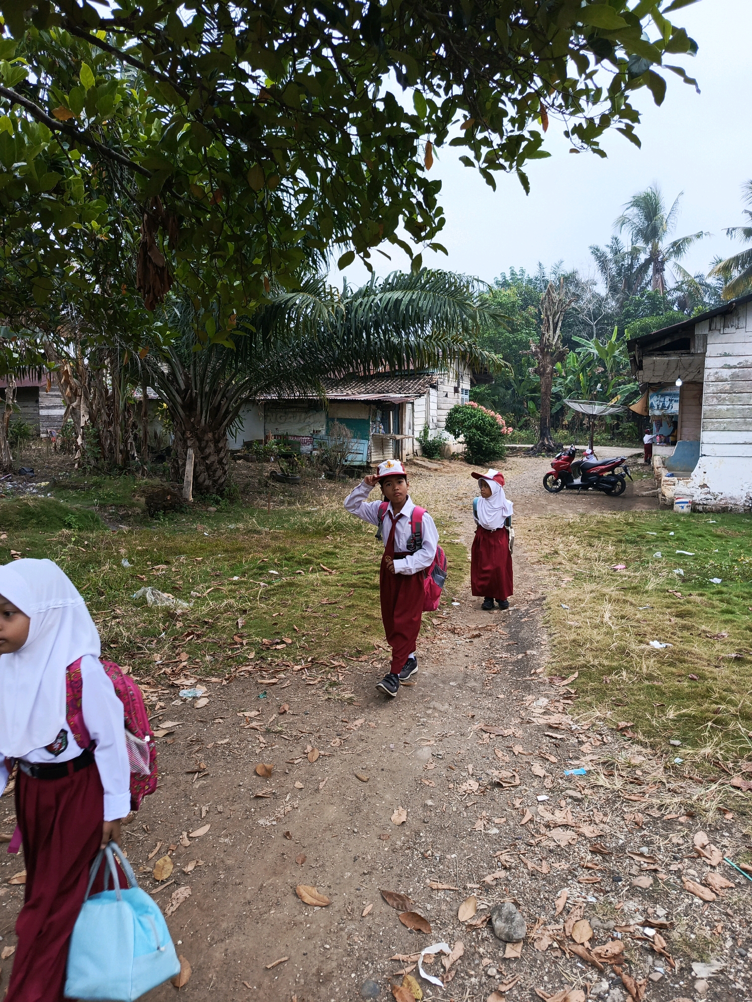 Three young students, two boys and one girl, are walking on a dirt path in front of simple buildings. The students are wearing school uniforms consisting of white shirts, maroon skirts or pants, and red hats. One boy in the foreground is saluting, while the girl in the background is wearing a white hijab. A red motorcycle is parked to the right of the path. The environment appears to be a rural or semi-rural area, possibly a village, with houses that have corrugated metal roofs and wooden or plank walls. Lush green trees and foliage are present throughout the scene, suggesting a tropical climate. The ground is covered with dirt, fallen leaves, and some scattered debris. The sky is overcast. The scene appears to be a typical day for students in Megang Sakti II, Indonesia.