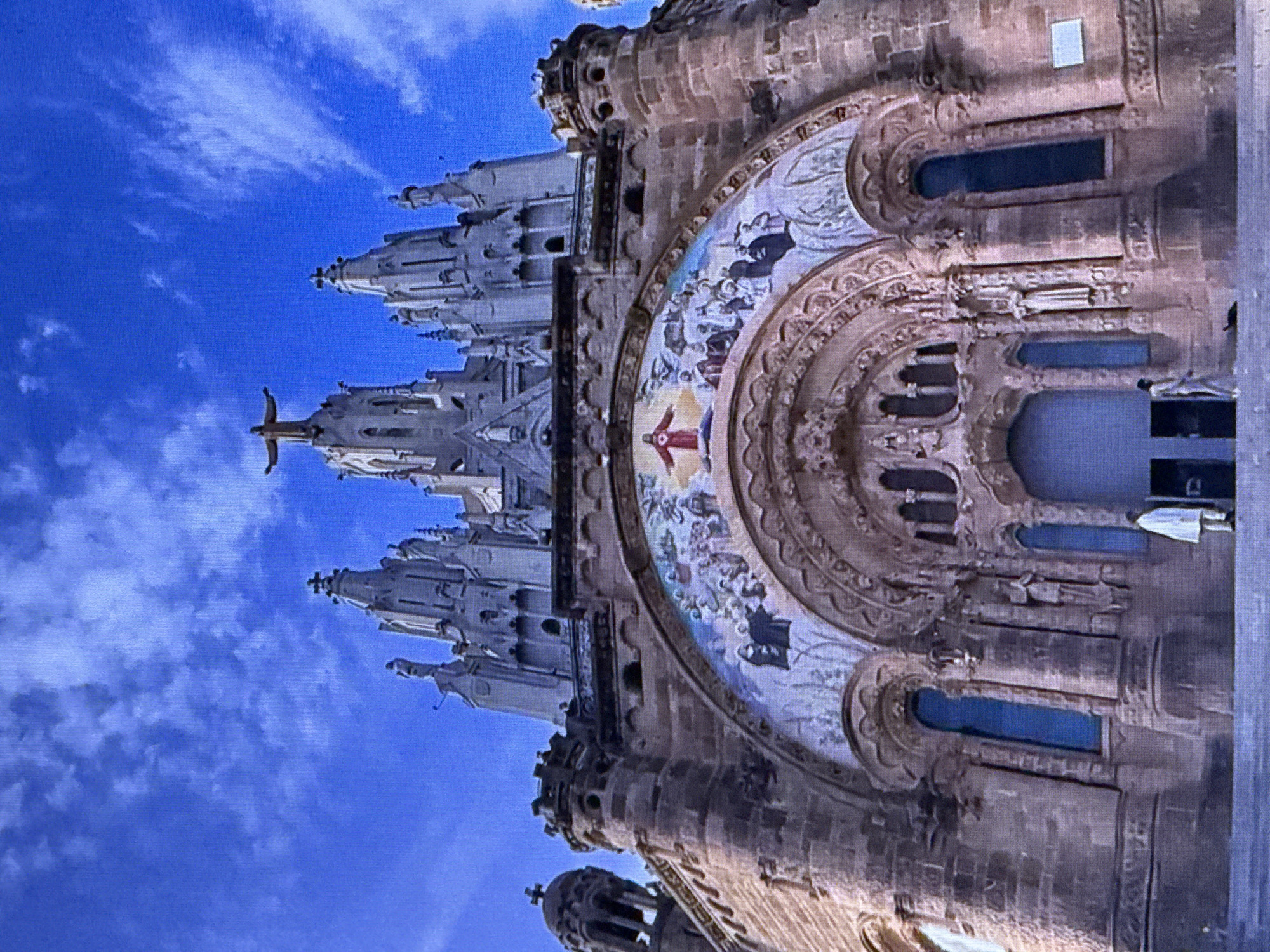 The media file captures a low-angle, close-up view of the exterior of a grand stone building, likely a church or cathedral, against a bright blue sky with scattered white clouds. The architecture is intricate and Gothic-inspired, featuring spires, turrets, and ornate carvings. A prominent circular mosaic adorns the facade, depicting figures in a religious scene, with a central image of a person in red with outstretched arms. Smaller figures are visible in the background of the mosaic.
The building's stone material appears weathered, suggesting age. The windows are dark, hinting at either interiors not lit or deep recesses. The scale of the structure is imposing, with detailed stonework emphasizing its grandeur. At the very top of one of the spires, a dark silhouette of a cross is visible. The lighting suggests it is daytime, possibly late morning or early afternoon, given the brightness of the sky. The overall impression is one of awe and architectural magnificence. No visible text is discernable. While the prompt states the location context is Dubai, United Arab Emirates, the visual cues within the image itself do not confirm or deny this specific location. The style of architecture is not typical of Dubai.