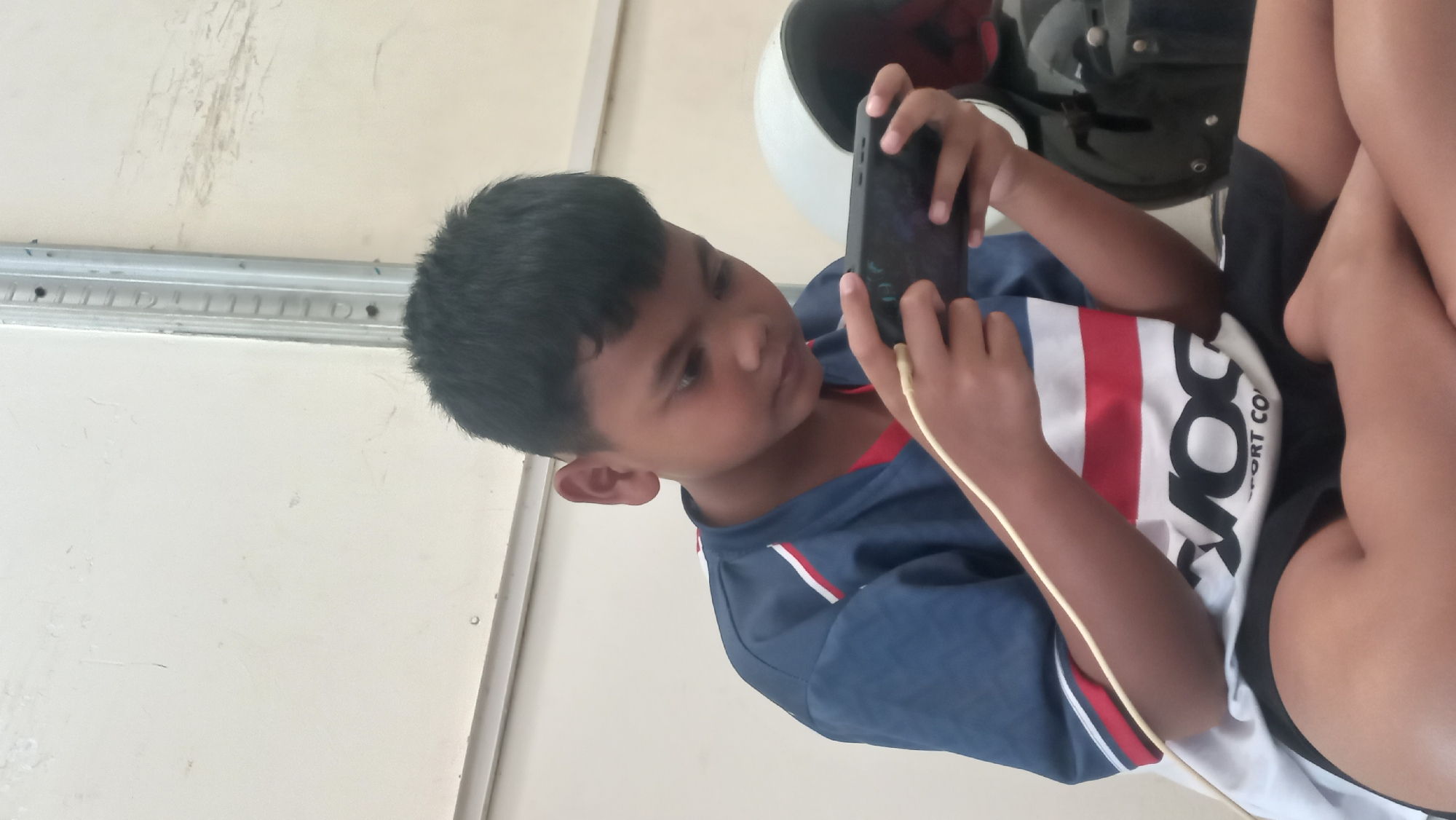 A young boy is sitting cross-legged, playing on a mobile phone. He is wearing a blue shirt with white and red stripes. A charging cable is plugged into the phone. In the background, a white helmet is visible, and the wall appears to be painted a light cream color. The scene takes place in Jetis Indah, Indonesia.