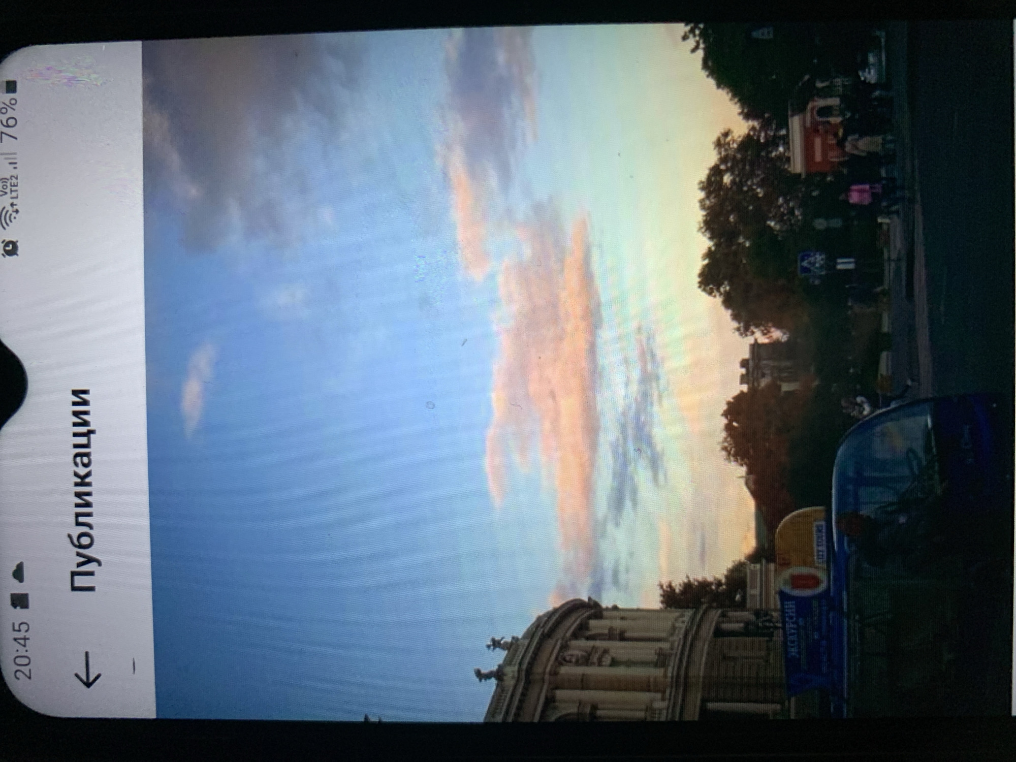 The image shows a smartphone screen displaying a photograph of a city street scene at sunset. The top of the screen shows the time as 20:45 and a battery indicator of 76%. The photograph itself features a wide, pale blue sky with scattered clouds tinged with pink and orange from the setting sun. In the foreground, a large, classical-style building with columns and statues is visible on the left side. A blue tour bus with "ЭКСКУРСИИ" (Excursions) written on it in white letters is parked in the street in front of the building. Several people are visible walking on the sidewalk and crossing the street. Trees line the street, and a traffic light and a pedestrian crossing sign are also present. The overall impression is of an urban environment during golden hour. The photograph is captured from a low angle, looking upwards towards the sky and the tops of the buildings and trees. The image does not provide any specific visual clues to confirm or deny the location as Lymanka, Ukraine.