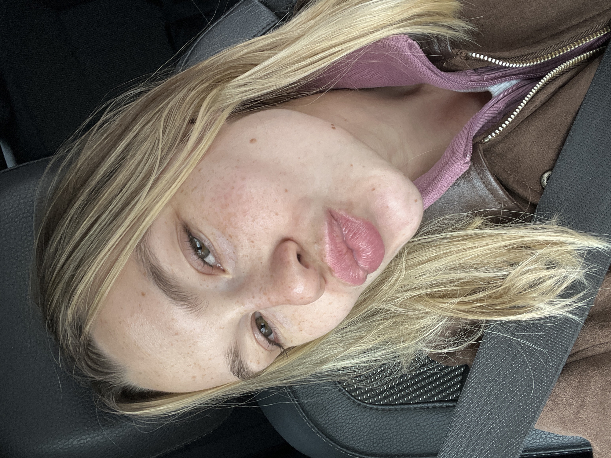 A close-up, slightly canted, selfie shot of a blonde woman in a car, wearing a seatbelt. She has fair skin with freckles, and her lips are full. She is wearing a brown jacket with a partially unzipped pink hoodie underneath. The interior of the car seat is black leather. The location of the image cannot be determined. There is no indication of the city of Rogachevka, Russia, in the image.