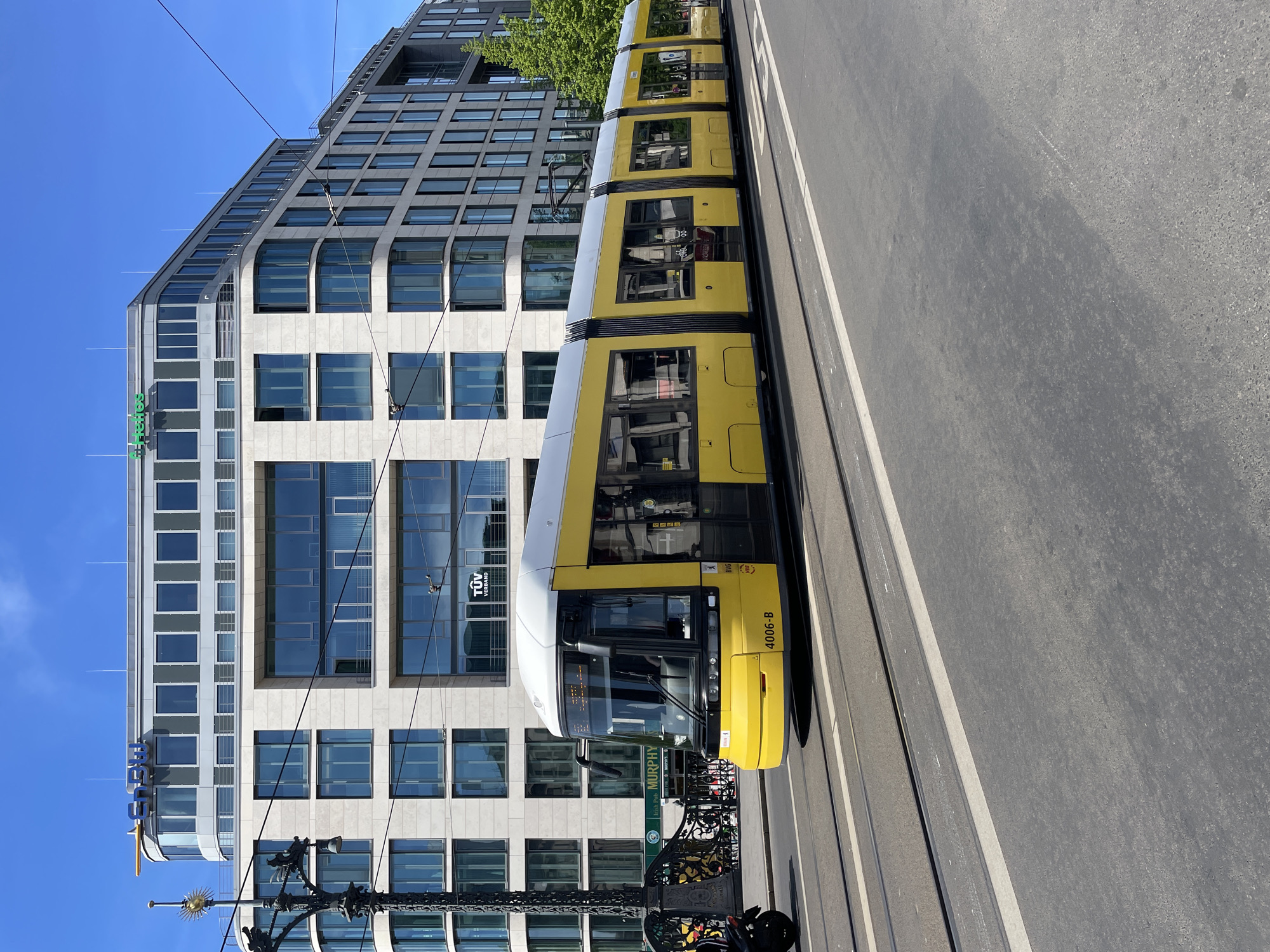 The image displays a bright, sunlit urban scene in Berlin, featuring a distinctive yellow tram alongside a modern architectural building. The tram, labeled "4006-B" on its front, occupies the right side of the frame, moving or stationary on its tracks. Its windows reflect the clear blue sky and parts of the building.
On the left, a large, multi-story building with a contemporary design features a facade composed of light-colored stone or concrete panels and numerous glass windows, some of which are rectangular, others slightly curved. Several corporate logos are visible on the building: a prominent green "SAP" logo near the top left, "TÜV VERBAND" visible through a tram window's reflection and on the building itself, and "MURPHY" on a sign on a lower floor. The building's upper sections have a unique, angular, and stepped design, with anti-bird spikes visible along its edges.
The clear blue sky indicates fair weather. Bright sunlight casts distinct shadows, suggesting it is daytime, likely mid-morning or afternoon. In the foreground, parts of an ornate black metal structure, possibly a historical lamppost or a decorative barrier, are visible. The ground consists of grey asphalt with white lines marking the tram tracks and road. The scene captures a typical, bustling environment within a modern European city.