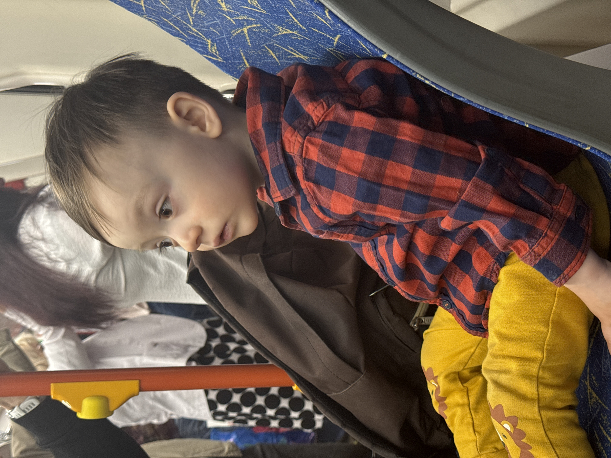 A young child is sitting on a vehicle seat with a blue and white patterned cover. The child is wearing a red and navy plaid shirt and yellow pants with a lion face design. The child is looking down and to the left. A brown coat hangs behind the child. In the background, a person with long brown hair is visible, along with other indiscernible objects. It is impossible to determine the location based on the image. The location could be Izhevsk, Russia, or another city.