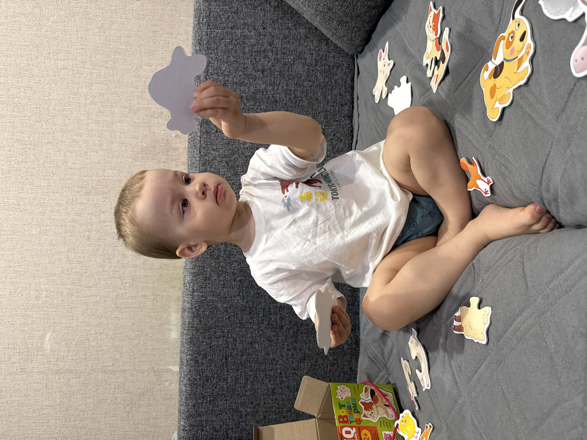A young child sits cross-legged on a gray couch, holding a white puzzle piece in their right hand. Other puzzle pieces depicting various animals, such as a dog, cat, fox, and sheep, are scattered on the couch around the child. The child wears a white t-shirt with a graphic design. To the lower left, a cardboard box with a puzzle visible inside is also present on the couch. The background includes a beige wall and the armrest of the gray couch. There is no information within the image that places the location in Rusanovo, Russia.