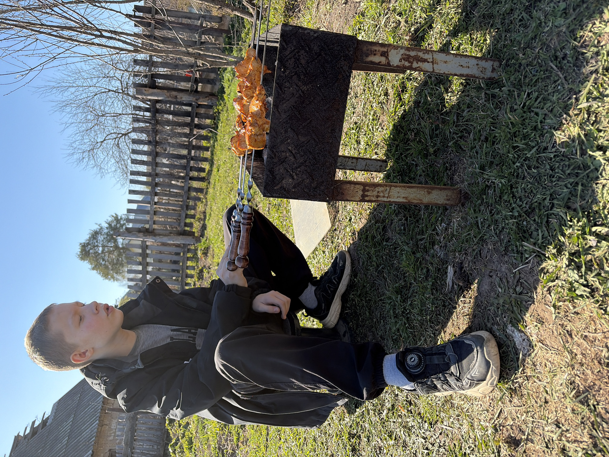 A young male sits on the ground, wearing black pants, a grey t-shirt, a black jacket, grey socks, and black sneakers. He is positioned next to a rusted metal barbecue grill on which skewers of cooking meat are placed. The location is an outdoor area in Чумна, Russia, with grass and a wooden fence in the background. The sky is clear and blue.