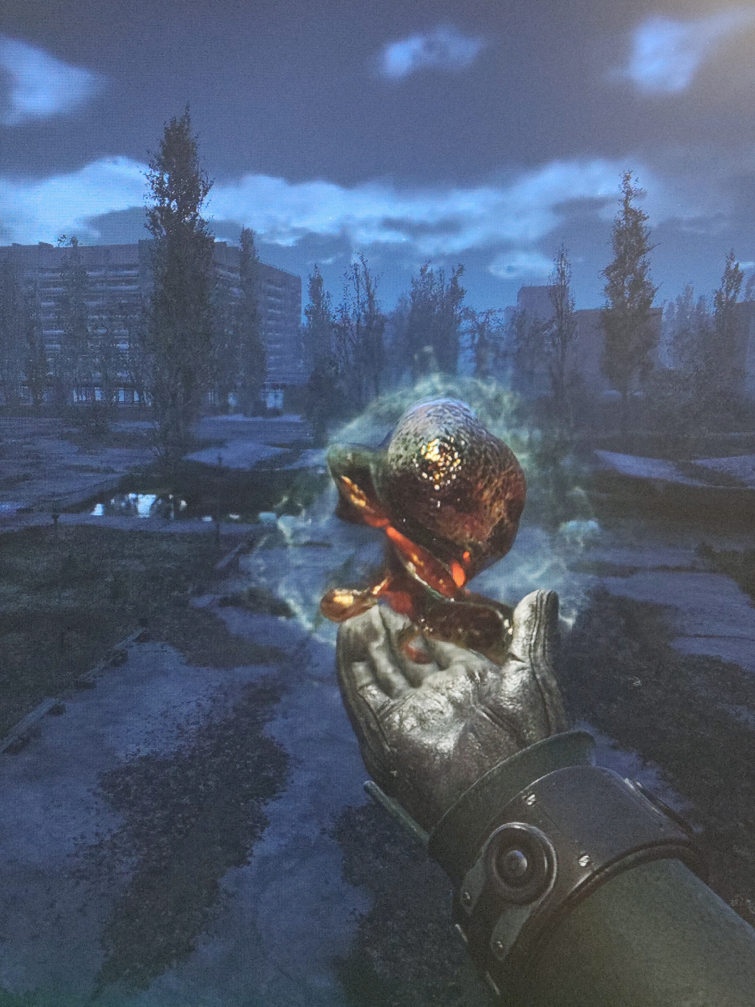 A first-person view depicts a gloved hand holding a luminous, organic object emitting a faint green glow. The background features a dilapidated urban landscape, characterized by derelict buildings and sparse vegetation under a cloudy sky. Puddles are visible on the ground. The image suggests an environment of decay and possible contamination.