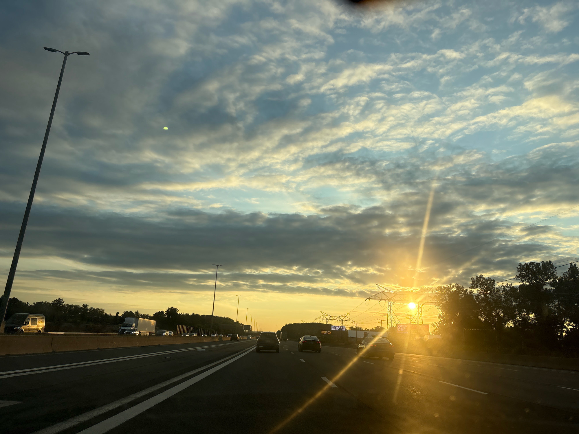 The image shows a highway scene in East Hartford, Connecticut, United States of America, during what appears to be sunset. The sun is low on the horizon, casting a warm glow and creating lens flares across the frame. Several cars and trucks are visible on the multi-lane highway, suggesting moderate traffic. Tall streetlights line the sides of the road, and power lines with large transmission towers are also present. The sky is filled with a mix of clouds, some dark and heavy, others lighter and illuminated by the setting sun, creating a dramatic sky. Trees and vegetation are visible beyond the highway. The overall impression is that of a typical evening commute on a highway under a dynamic sky.