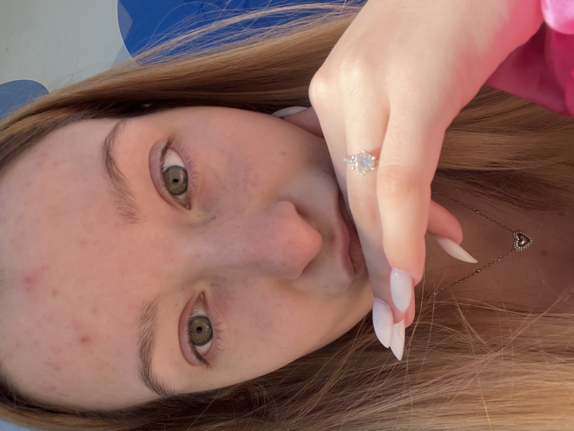 A close-up selfie photograph depicts a light-skinned person with brown hair and green eyes. The person's face exhibits freckles and minor skin blemishes. Their hand, adorned with a silver ring featuring a prominent stone, partially obscures their mouth. Artificial, long white nails are visible on the fingers. A silver necklace with a heart pendant rests against the person's neck. The background features a blur of colors including blue, white, and pink, suggesting an indistinct environment. No information regarding the location in Varna, Bulgaria is discernible from this image.