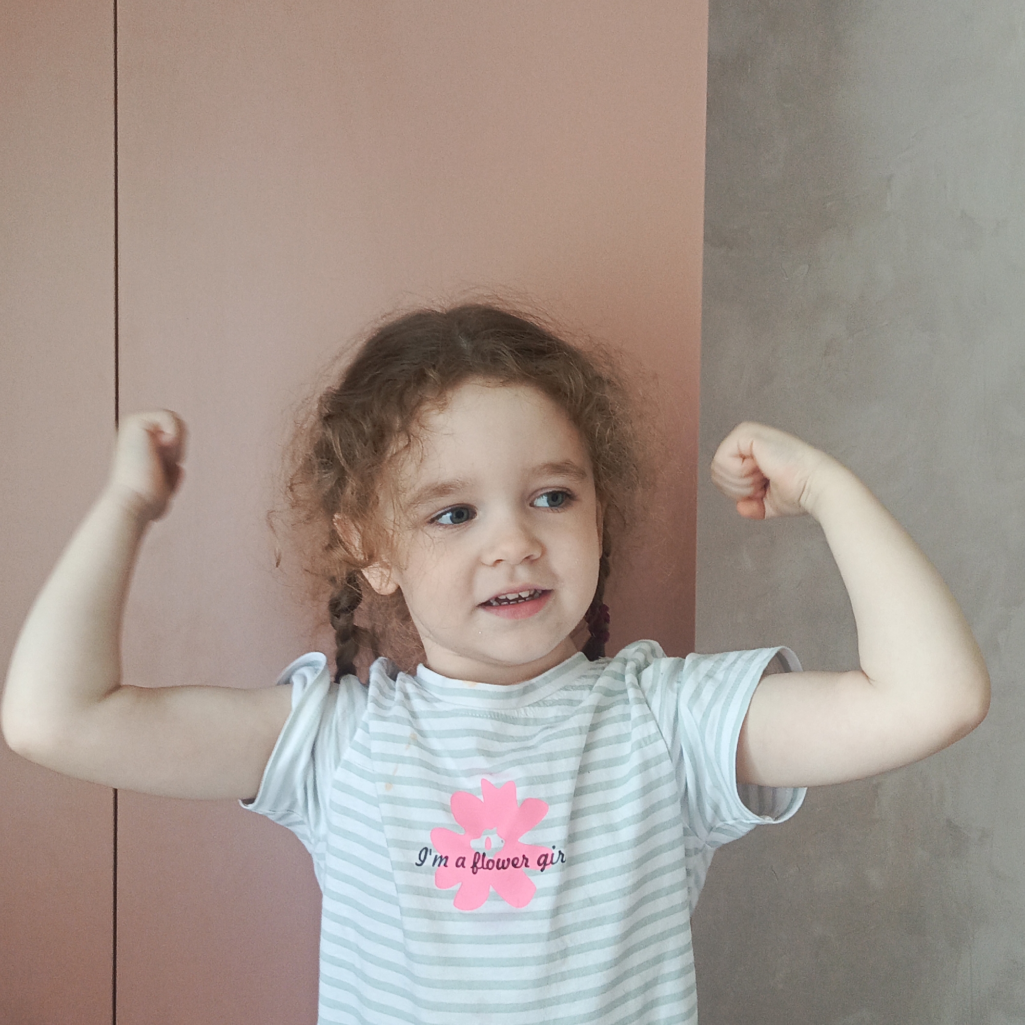 A young girl with curly brown hair and two braids is standing indoors and flexing her biceps. She is wearing a light blue and white striped t-shirt with a pink flower graphic and the text "I'm a flower girl" printed on it. The girl is looking to her right with a slight smile. Behind her, the background is a split between a soft pink wall and a textured grey wall. There are no objects or people suggesting a specific location in Марковский, Russia.