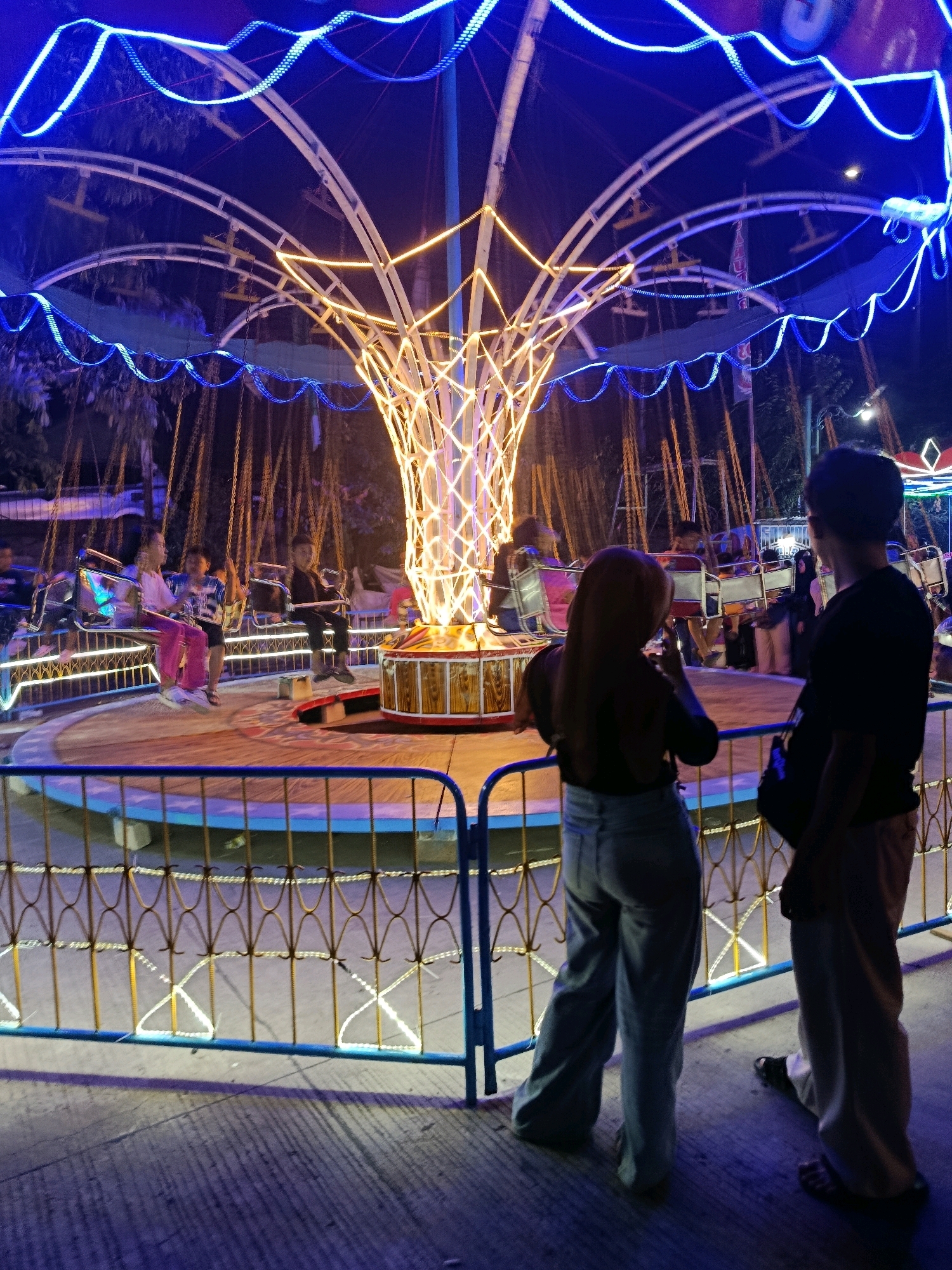 The image depicts an amusement ride, likely a swing ride, illuminated by blue and yellow lights at night. The ride features a central structure with a cage-like design, from which numerous chains hang, each suspending a seat. Several people are seated on the ride, with some appearing to be children and others adults. In the foreground, two individuals are standing behind a decorative fence. One is a person with a red hijab and light-colored pants, facing away from the camera and towards the ride. The other is a person in a black t-shirt and light-colored pants, also facing the ride. The ground is paved, and the overall setting suggests an outdoor entertainment area. The presence of trees in the background indicates an urban or suburban environment. The sign "KARNAVAL MALAM BALARAJA" visible in the background confirms the location as Balaraja, Indonesia.