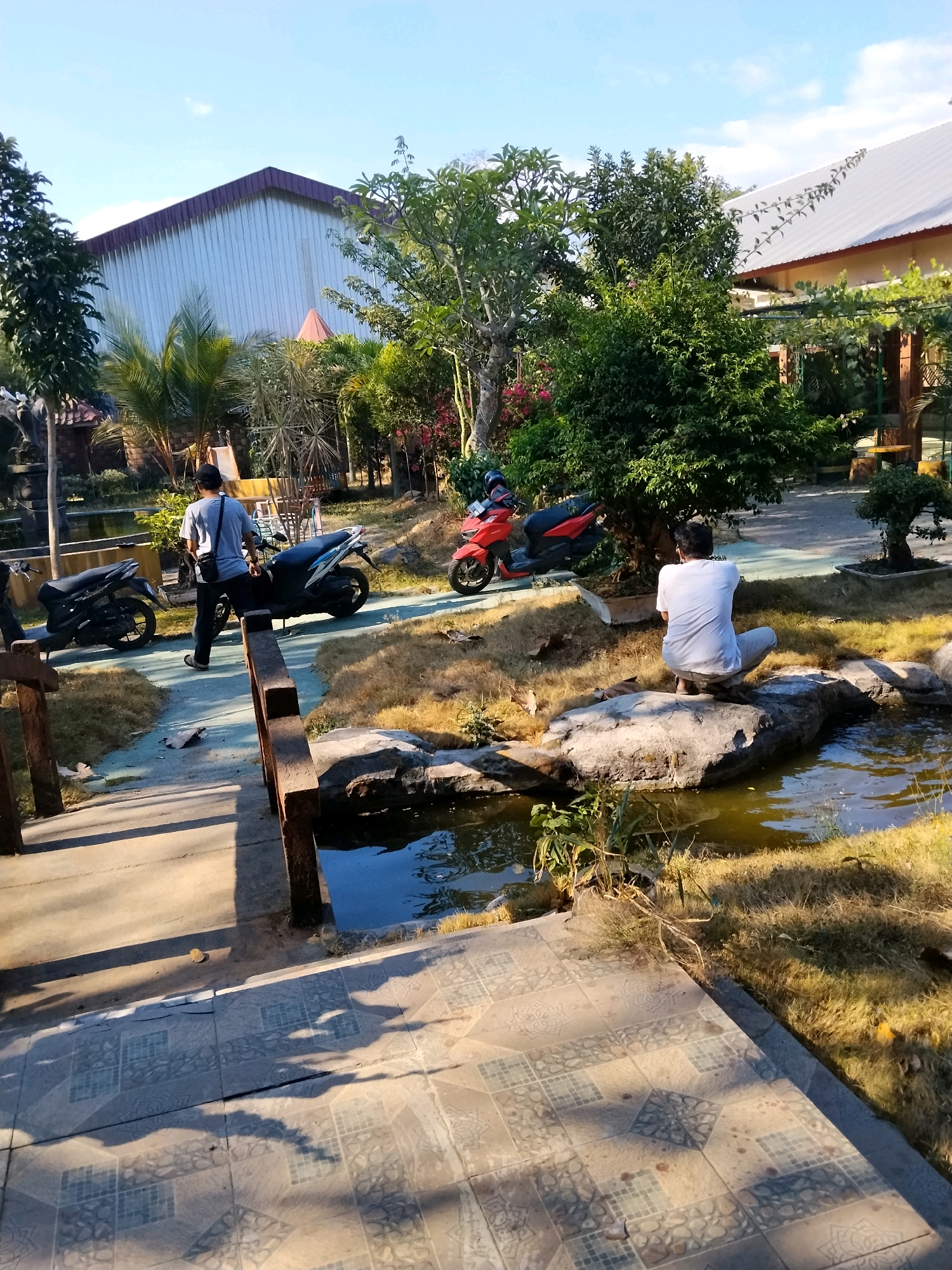 The image depicts an outdoor scene at what appears to be a recreational area or park in Pekalongan, Indonesia. In the foreground, a tiled pathway casts long shadows from a wooden bridge structure. Beyond the bridge, a shallow pond or water feature is visible, bordered by rocks and dry grass.
Two individuals are present. One man, wearing a light-colored t-shirt and dark pants, is walking away from the camera towards a blue and black motorcycle parked on a paved path. He has a dark bag slung over his shoulder. Another person, wearing a white t-shirt and light-colored pants, is seated on a large rock next to the water feature, facing away from the camera, appearing to be looking into the water or at something beyond it.
Several motorcycles are parked in the scene: a black motorcycle on the left, a blue and black motorcycle in the middle, and a red motorcycle on the right, partially obscured by trees. In the background, there are buildings with distinctive roofs, one appearing to be a large warehouse-like structure with a corrugated metal facade and a dark-colored pitched roof, and another building with a lighter-colored pitched roof. Lush green trees and smaller plants are scattered throughout the environment, some appearing to be mature trees and others more like decorative bushes or bonsais. The sky is blue with a few scattered clouds, suggesting a clear day. The overall impression is of a sunny day in a semi-urban or semi-rural setting.